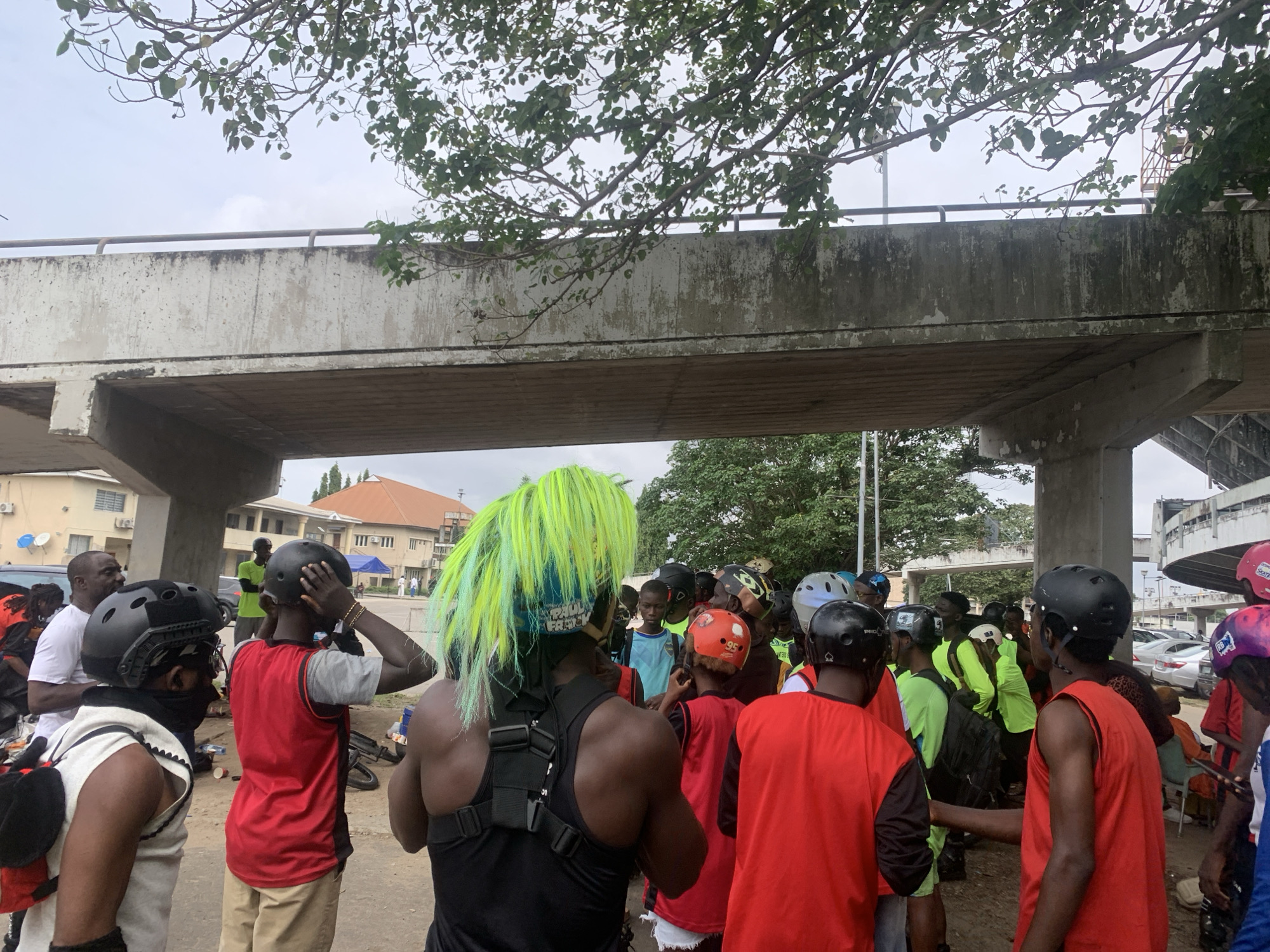 A group of young people wearing protective gear, including helmets, are gathered under an overpass in what appears to be an urban environment. The individuals are predominantly dressed in casual athletic wear, with some wearing bright colors. A person in the foreground on the left is wearing a white vest over a black garment and a black helmet. To their right, another person wears a red sleeveless shirt and a black helmet. In the center, a person with bright green and yellow spiky hair, also wearing a helmet, stands out. The group seems to be preparing for or resting from an activity, possibly rollerblading or skateboarding, as evidenced by the protective gear and a backpack visible. The background shows buildings, parked cars, and lush green trees, suggesting a tropical or sub-tropical climate. The overpass is a prominent feature of the scene, casting shadows on the group below. The overall atmosphere appears casual and communal, with individuals engaged in conversation or observing their surroundings. The scene takes place outdoors, with an overcast sky.
