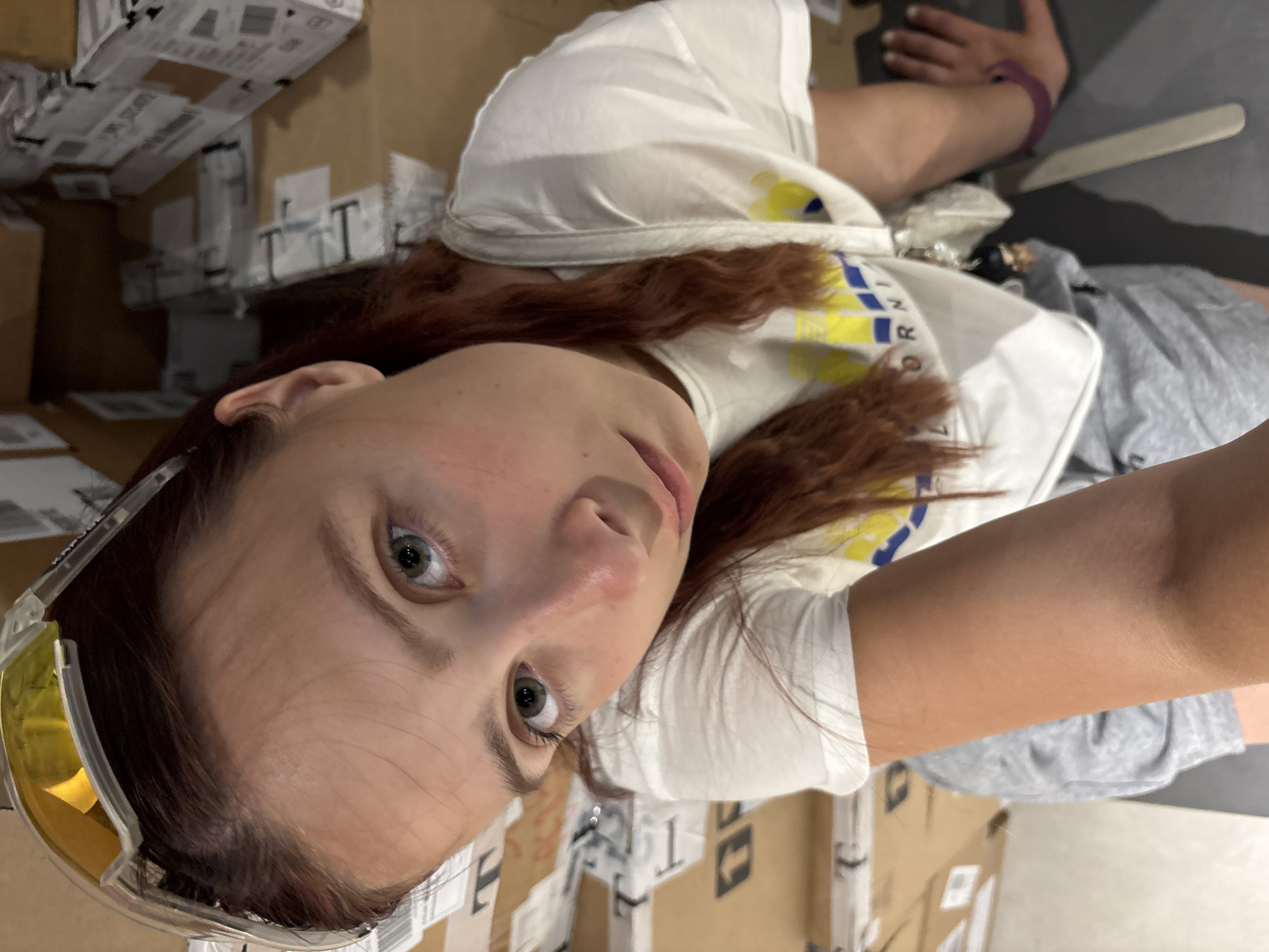 In Izhevsk, Russia, a young woman with reddish-brown hair and green eyes is captured in a selfie-style photograph. She is wearing a white t-shirt with a yellow and blue graphic print and grey shorts. Yellow-tinted sports sunglasses are perched on her head. The background is filled with stacks of cardboard boxes, many of which appear to be shipping or packaging materials. One box has visible text that includes "ORN" and "RNI", suggesting a brand name or labeling. The overall setting suggests an indoor environment, possibly a warehouse, storage area, or a room filled with merchandise. The lighting appears to be natural or ambient, casting soft shadows. The woman's right arm is visible, and she is wearing a purple bracelet. A thin, flat object, possibly a ruler or a piece of trim, lies on the floor to her right. The image is taken from a high angle, looking down at the woman.