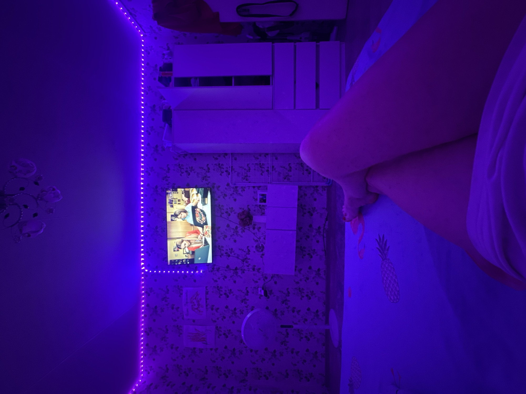 A bedroom interior in Chaikovsky, Russia, illuminated with purple LED strip lights, displays floral wallpaper, a television showing a program, a white dresser, a white fan, and pineapple-patterned bedding, with a person's legs visible in the bed.