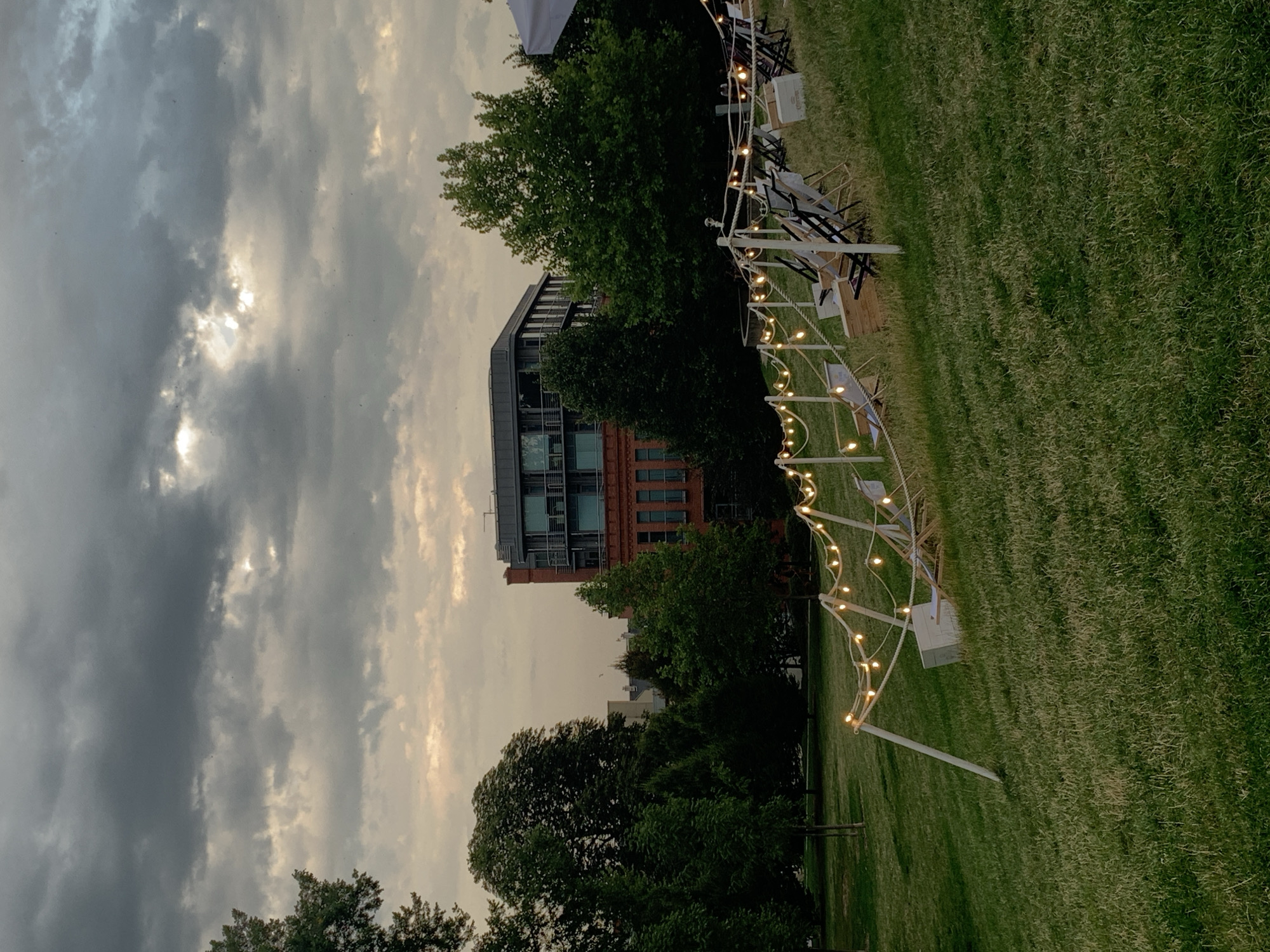 The image captures an outdoor seating area on a grassy slope, set against a backdrop of trees and buildings in Poznan, Poland. The sky is overcast with dramatic grey clouds, with patches of lighter sky visible through breaks. A string of globe lights runs along the top of the slope, supported by white poles, creating a festive atmosphere. Several deck chairs are arranged on the grass, some appearing to be made of canvas and wood, suggesting a place for relaxation or an outdoor event. Behind the chairs, a white tent or canopy is partially visible on the right. In the mid-ground, several lush green trees are prominent, and behind them, a distinctive red brick building with modern glass windows stands out. Further in the background, more buildings are visible, indicating an urban setting. The overall scene suggests a tranquil evening gathering or an area designed for outdoor leisure in Poznan.