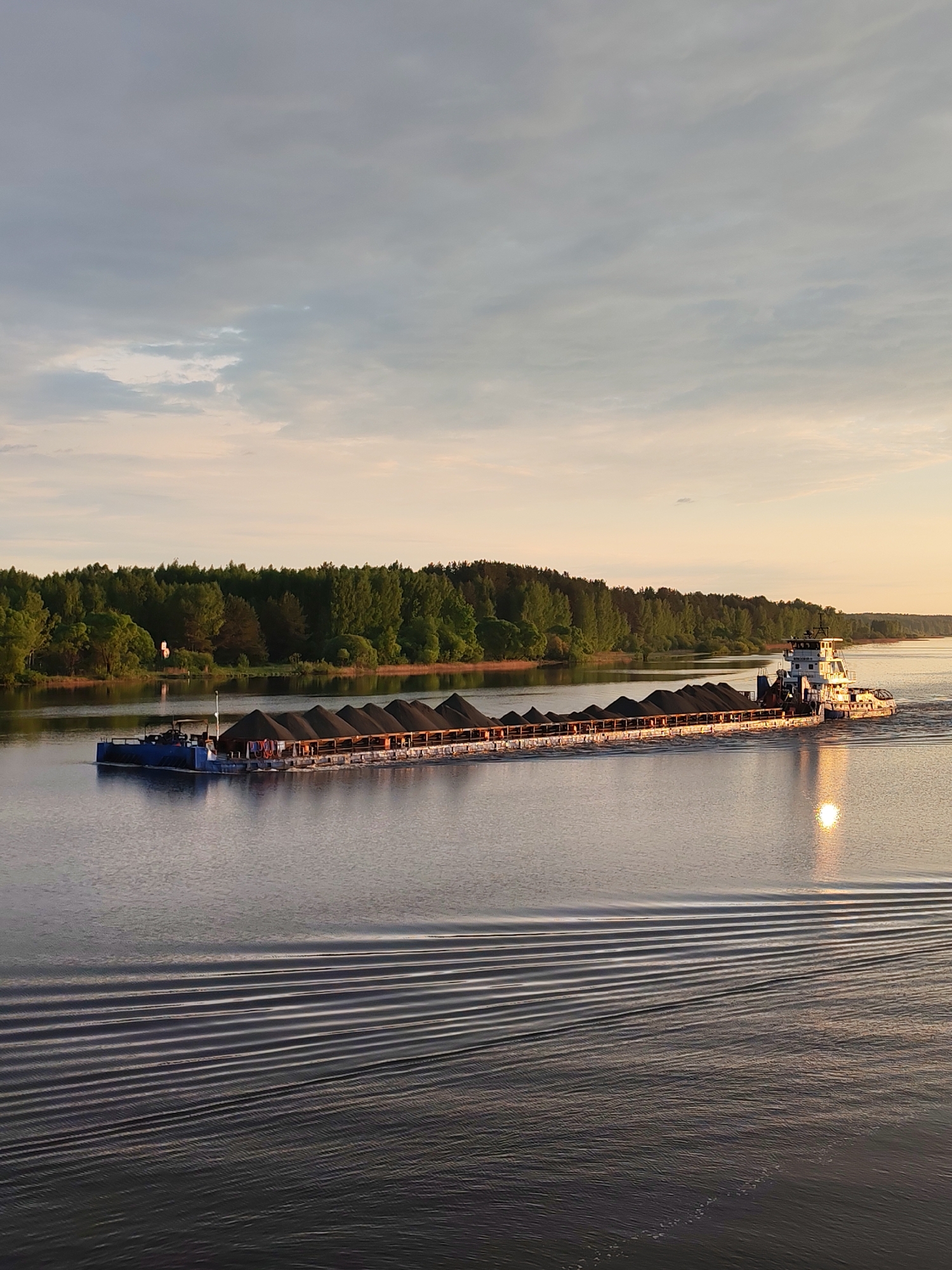A tugboat towing a barge laden with dark material traverses a river under a sky with varying cloud cover and hints of sunset. The tugboat is white with a visible cabin structure. The barge is long and flat, seemingly filled with conical piles of the dark material. The river's surface is rippled, with a sun reflection visible. A forested shoreline borders the river in the background. The location is unknown.
