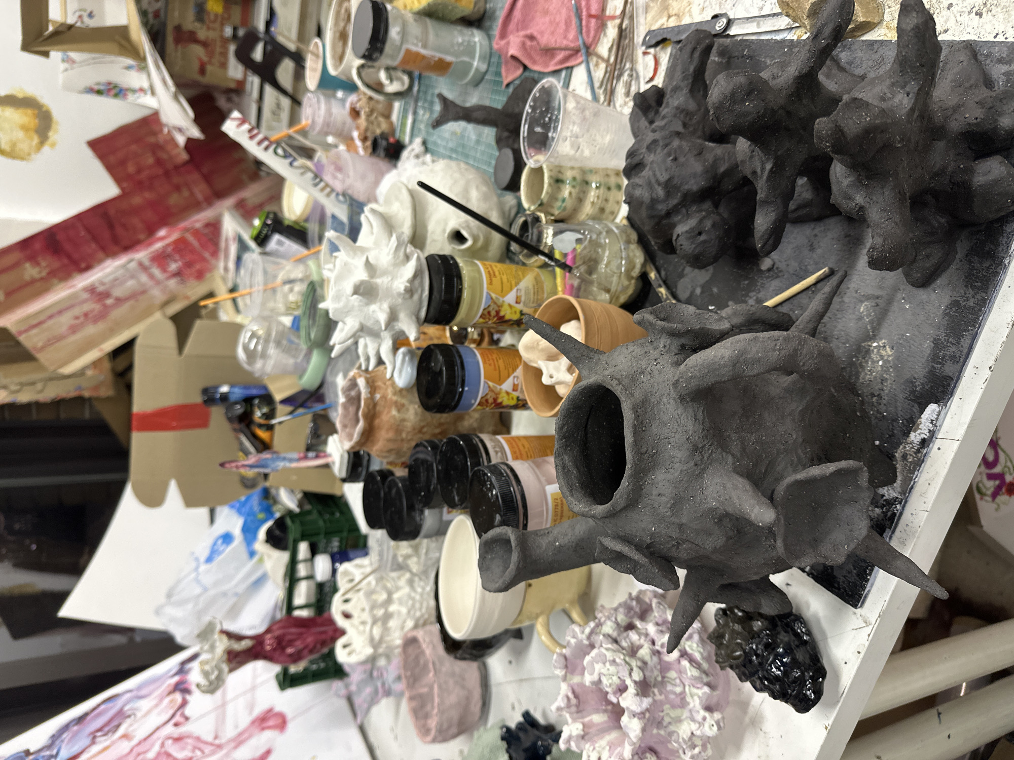 The image depicts a cluttered art studio environment with a variety of art supplies and sculptural pieces. In the foreground, there are several dark-colored, textured sculptures, possibly made of clay or a similar material, resting on what appears to be a painted surface or worktable.
The background features a jumble of art-related items including jars, paint bottles, plastic cups, and paper. There are also glimpses of paintings or works on paper, some with red and white coloring. The environment is disorganized and suggests a working artist's space.
No specific location in Moscow, Russia is discernible from the visual information provided.