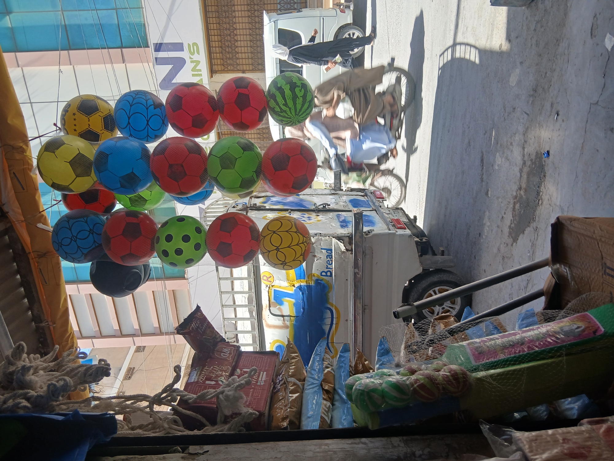 The image captures a street scene in Quetta City Tehsil, Pakistan. A vendor displays a variety of brightly colored soccer ball-shaped balloons, alongside other merchandise including packaged snacks and toys, likely from a makeshift stall. A three-wheeled vehicle is visible, parked in the background. In the distance, blurry figures of pedestrians and a cyclist can be observed. Buildings with metal structures and awnings surround the scene.