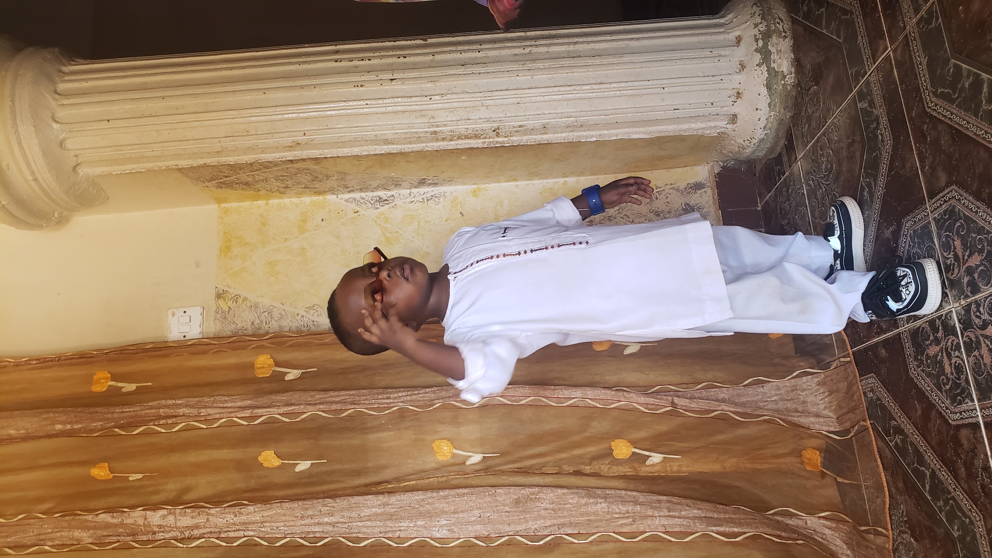 A child, dressed in a white robe-like garment with a decorated neckline and wearing black and white sneakers, lies horizontally on a decorated brown fabric placed over tiled flooring. The child also has sunglasses on their head and a blue bracelet on their left wrist. The flooring is patterned in a dark brown design. A wall with peeling paint and a white decorative molding sits above the child. A portion of a brown curtain with yellow floral designs is visible on the left. The location is presumably Narayi, Nigeria, based on the prompt.