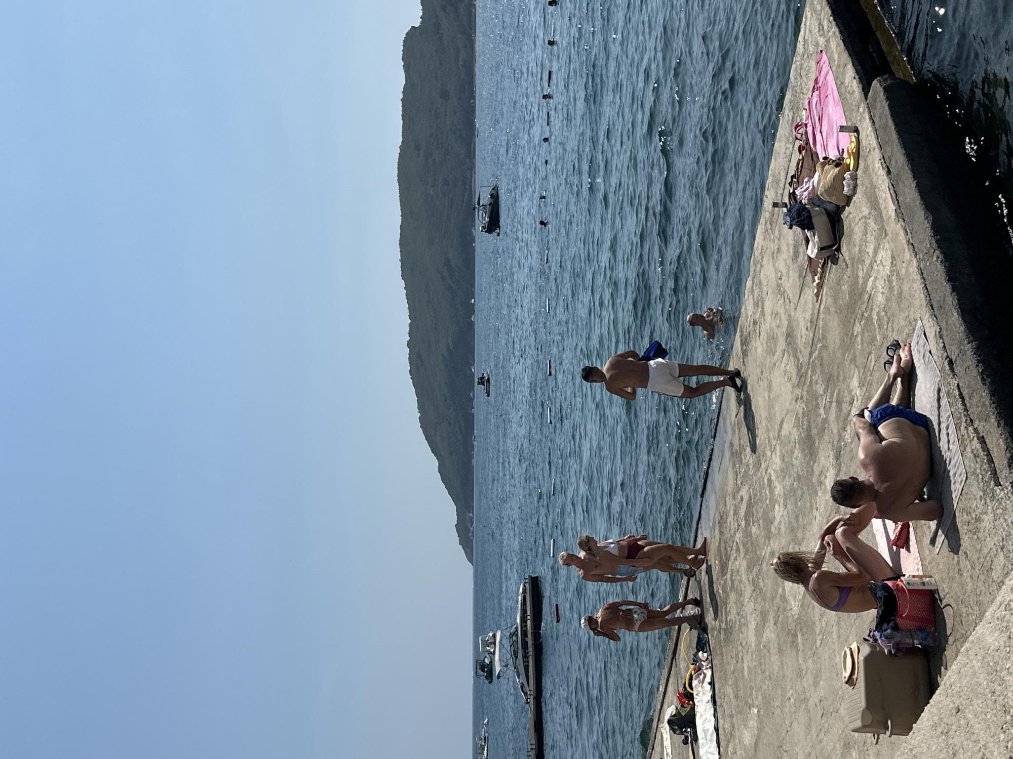 A sunny day at the Mogren beach in Budva, Montenegro, with a clear blue sky and calm sea. Several people are present, either relaxing on the concrete pier or swimming in the water.
On the pier, a couple sits on towels, the woman in a purple bikini and the man in blue swim trunks. Nearby, a group of four individuals stands, two women in bikinis and two men, one in swim trunks and the other in shorts. Another man in white shorts stands at the edge of the pier, facing the water. Further along the pier, a collection of towels, bags, and flip-flops is visible.
In the sea, several people are swimming or wading, with buoys marking a designated area. Various boats are anchored or moored in the distance, including a large white yacht near a pier on the left side of the image. A green hill covered in trees forms the backdrop, characteristic of the Budva coastline. The concrete pier extends into the water, providing a vantage point for beachgoers to enjoy the scenic surroundings.