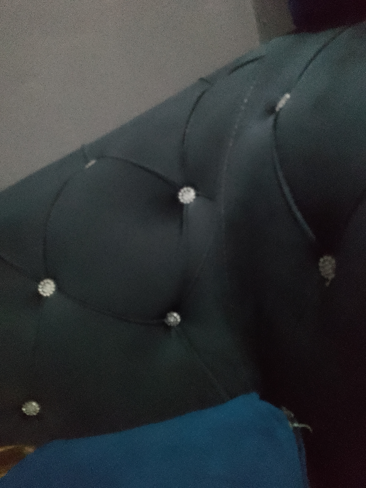 The image shows a close-up, angled view of a dark teal, tufted upholstered surface with diamond-like embellishments. The surface appears to be part of a piece of furniture, possibly a sofa or headboard, given the deep buttoning and the soft material. In the foreground, a section of bright blue fabric is visible, suggesting it is another item of furniture or textile placed in front of or below the upholstered surface. The lighting is dim, casting shadows and highlighting the texture of the upholstery. The background is out of focus and appears to be a plain, textured wall, possibly in a neutral gray tone. There is no visible indication of the city of Jauro Yinu, Nigeria, or any context related to its location. The image solely focuses on the details of the upholstered furniture and the blue fabric.
