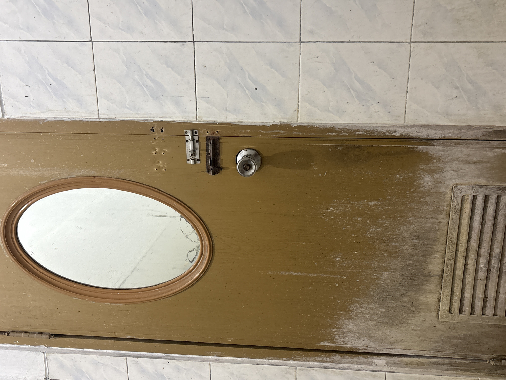 A worn, brown wooden door stands in a tiled room. The door has an oval mirror with a wooden frame set into its upper half. A silver doorknob is positioned below the mirror. Two metal latches, one appearing rusted, are mounted on the door to the right of the doorknob. The lower portion of the door features a decorative vent. The wall to the right of the door is covered in white tiles. The floor is not visible in the frame. The overall scene suggests an interior space, potentially a bathroom or utility room, in a building located in Palimanan, Indonesia, showing signs of age and use.