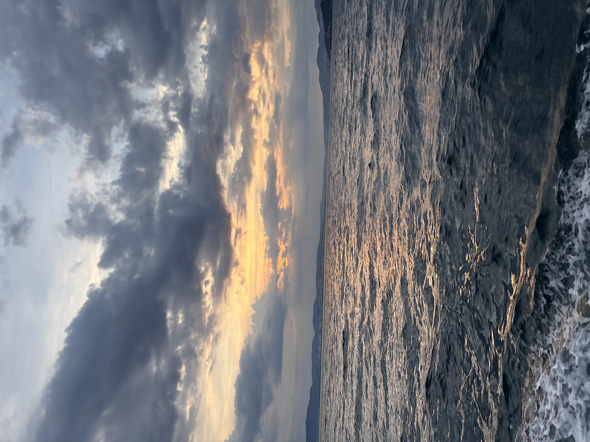 A seascape at sunset shows a body of water with choppy waves and a faint trail of white foam in the foreground. In the distance, a hazy, dark shoreline with hills and some buildings is visible under a dramatic sky filled with dark grey and bright orange clouds. The sun's rays filter through the clouds, casting a warm glow on the water's surface. The location is identified as Kabardinka, Russia.