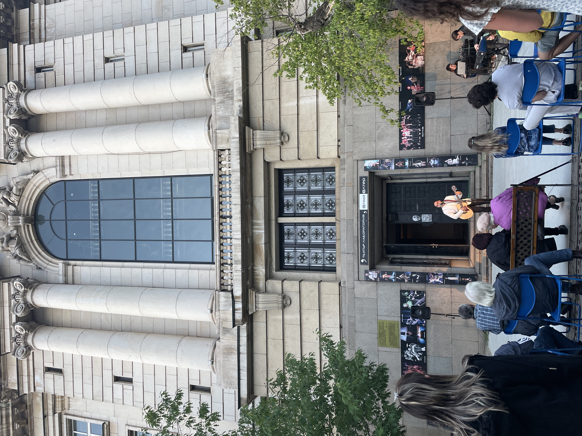This image depicts an outdoor musical performance taking place in front of a grand, classical stone building in Sofia, Bulgaria. The building features imposing, light-colored stone architecture with large, fluted columns topped with ornate capitals, an arched window, and decorative wrought-iron grates on other windows.
In the center of the scene, a male musician, wearing a light-colored shirt, is actively playing an acoustic guitar and singing. He is positioned near the main entrance of the building, which is adorned with several large posters showcasing images of theatrical or dance performances. An audience is seated on blue folding chairs, facing the performer, attentively watching the event. Other individuals are also visible, some standing or partially obscured, observing the performance. A speaker on a stand is part of the musical setup. Further back, another group of musicians with a keyboard and other equipment can be seen, suggesting a larger event or multiple acts.
The natural lighting suggests it is late afternoon or early evening, with clear weather conditions. Green foliage from surrounding trees is visible, adding to the outdoor setting. Cyrillic text visible above the entrance reads "ТЕАТЪР ЛАБОРАТОРИЯ," which translates to "Theater Laboratory," identifying the building as a cultural venue. A QR code is also visible near the door, and a plaque is mounted on the wall near the speaker, though its text is illegible. The overall mood appears calm and appreciative, with the audience engaged in the live music.
