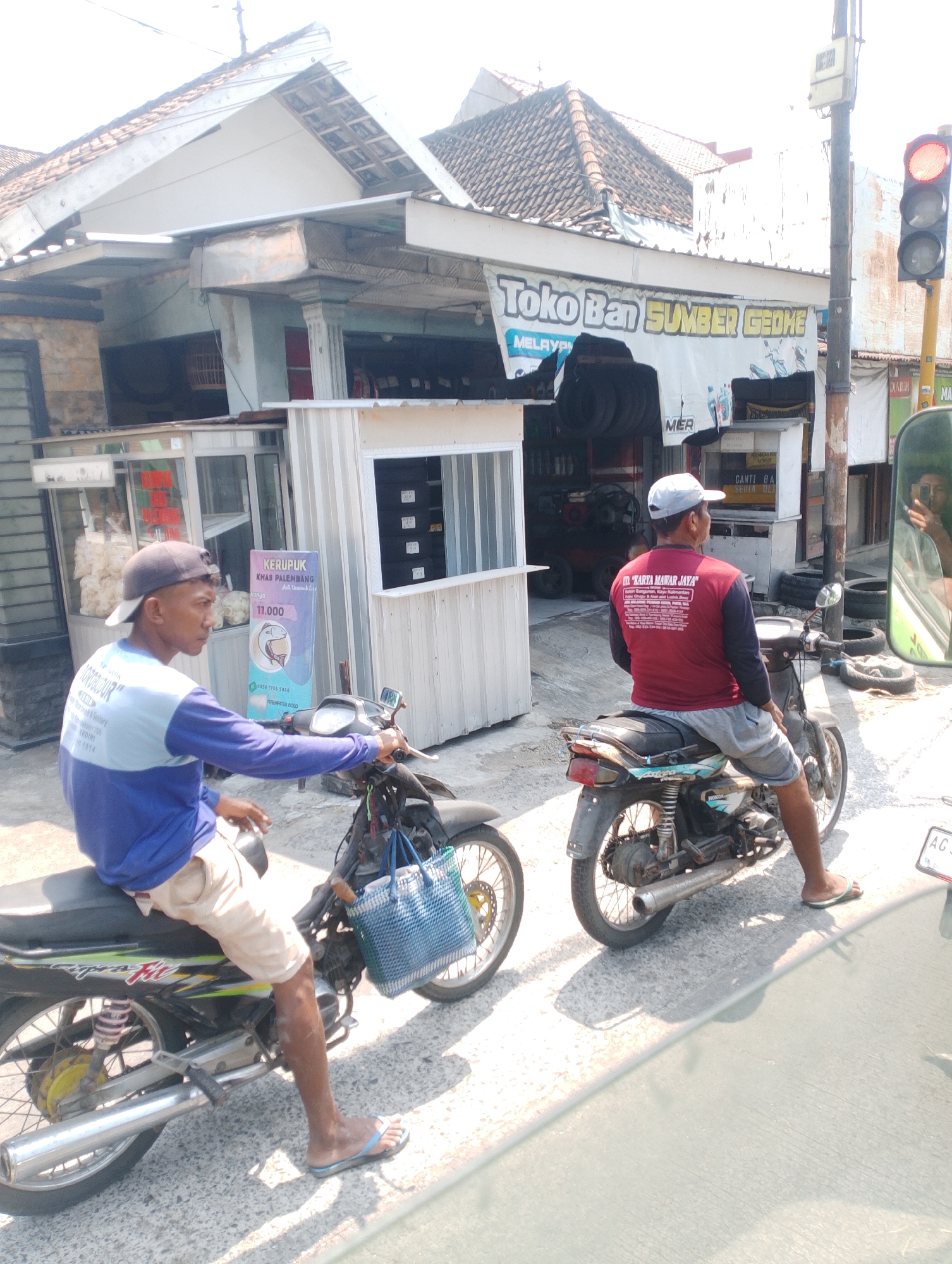 Two men are on motorcycles in what appears to be a street scene in Plemahan, Indonesia. The man on the left is wearing a blue and light blue long-sleeved shirt and light-colored shorts, with a dark baseball cap. He is seated on a dark-colored motorcycle and looking to his right. Attached to the front of his motorcycle is a blue woven bag. The man on the right is wearing a red long-sleeved shirt with white text on the back, light gray shorts, and a light-colored cap. He is seated on a dark-colored motorcycle and looking to his left. Both men are wearing flip-flops.
In the background, there are several buildings. One building has a sign that reads "Toko Ban SUMBER GEOHE" and appears to be a tire shop, with stacks of tires visible. Another building has a display of packaged goods, possibly snacks, and a sign for "KERUPUK PALEMBANG". A traffic light with a red signal illuminated is visible on a pole to the right of the frame. The ground is paved with asphalt and shows shadows cast by the sun. The overall environment suggests a commercial area on a sunny day.