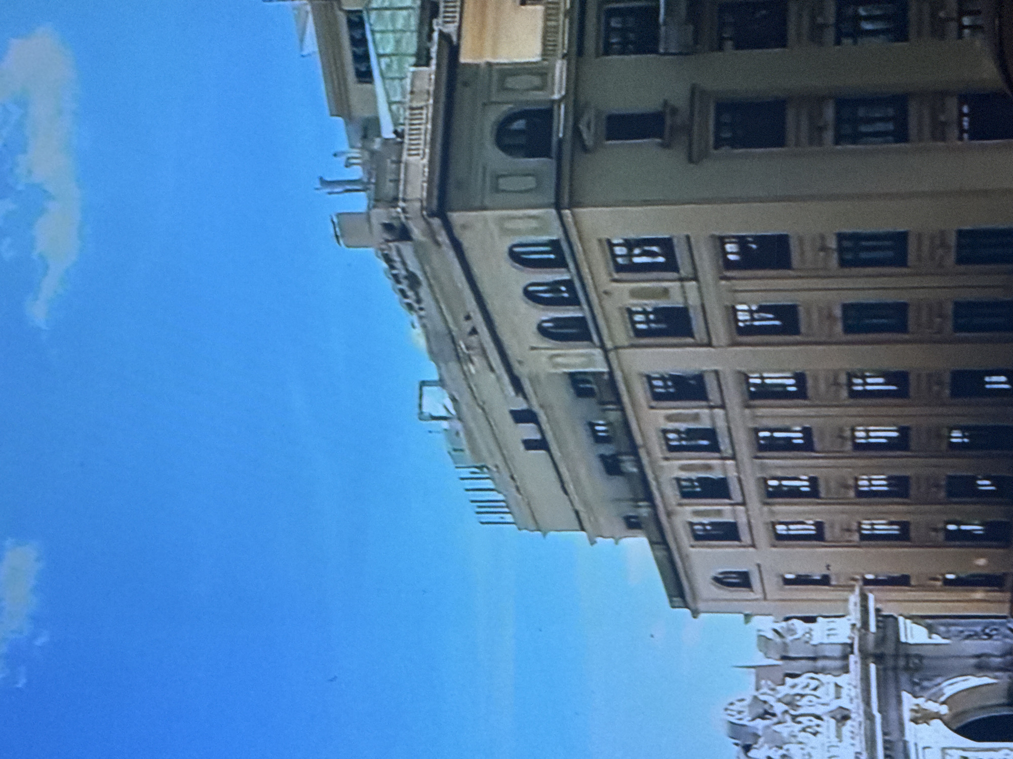 A high-angle, tilted shot depicts a beige building under a bright blue sky with faint clouds. The building is multi-storied with numerous windows and ornate architectural details. Part of a more decorated structure is visible at the bottom right, suggesting a street-level detail. The location is Moscow, Russia.