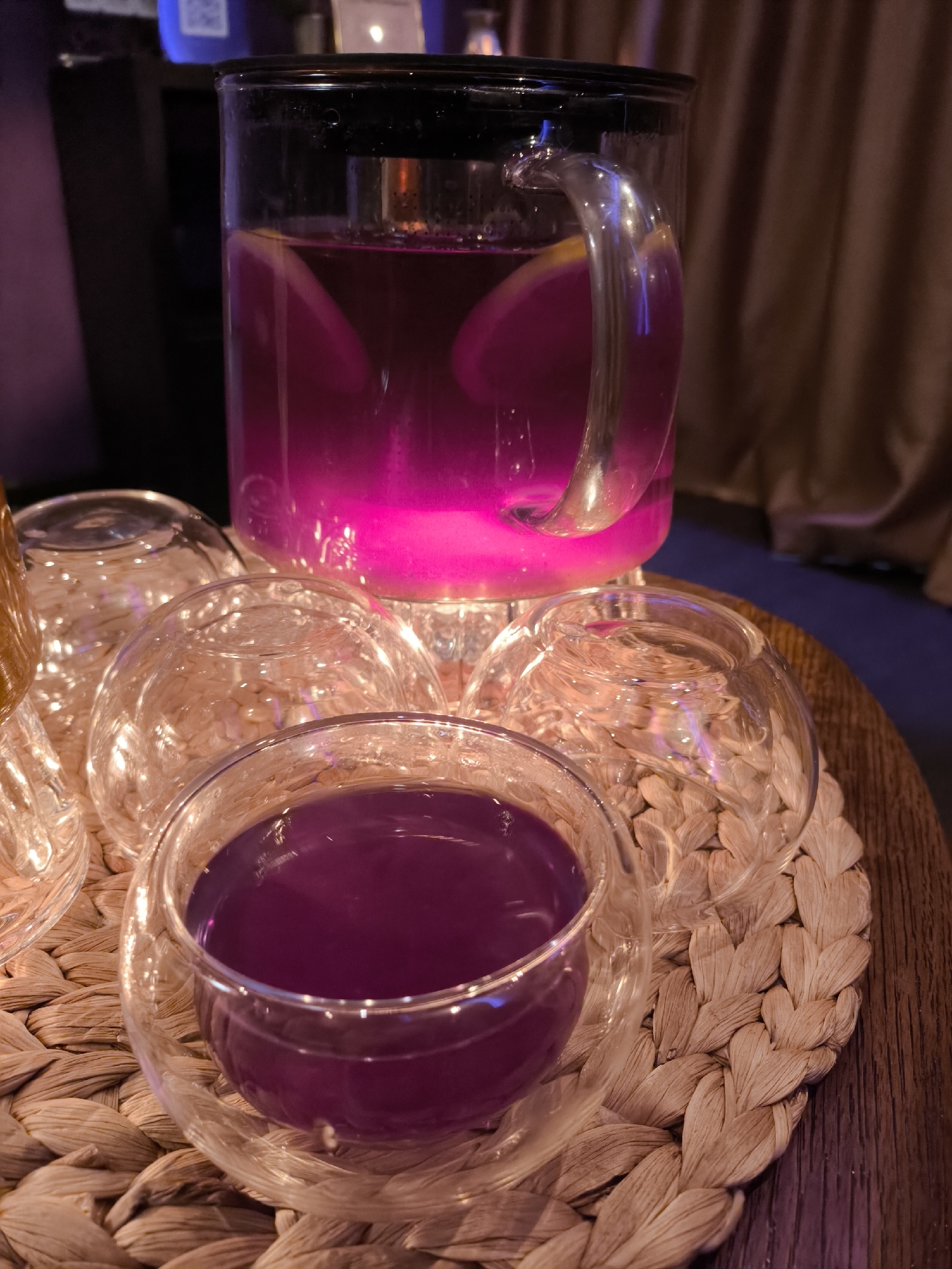 A medium-shot presents a beverage service atop a woven mat, likely situated indoors. A clear, glass pitcher, filled with a purple-hued liquid and slices of what appears to be lemon, is positioned at the back. Below the pitcher are multiple clear, glass cups, one of which is filled with the same purple liquid. The woven mat rests on a dark wooden surface. The scene's context might involve a beverage presentation or tasting, though there are no identifiable individuals, specific actions, or interactions present. The location is unknown.