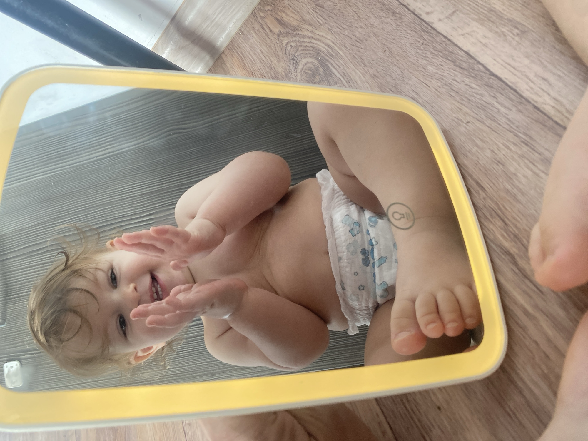 A baby wearing a diaper is reflected in a yellow-bordered mirror. The baby is smiling and has their hands raised. The background appears to be a wood-patterned floor. The location is unknown.