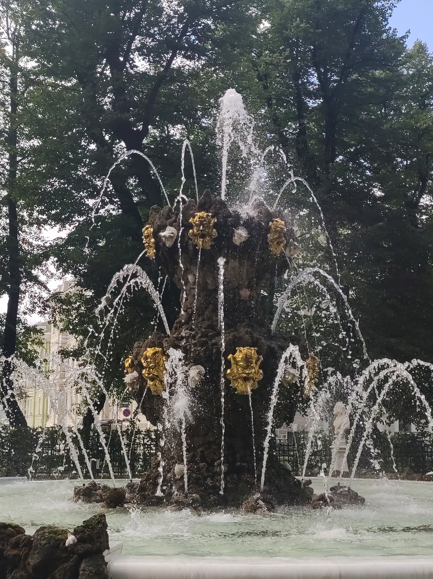 The image shows a fountain with water spraying upwards and outwards, surrounded by trees. The fountain has a central structure made of dark, rocky material, adorned with golden, grotesque faces that appear to be spouting water. Several streams of water arc and fall into the basin below. In the background, through the trees and water spray, a pale building with many windows can be seen. To the right of the fountain, a white statue of a bust on a pedestal is partially visible. The overall scene is of an outdoor garden or park area. This is the Samovar Fountain located in the Alexander Garden in Saint Petersburg, Russia.