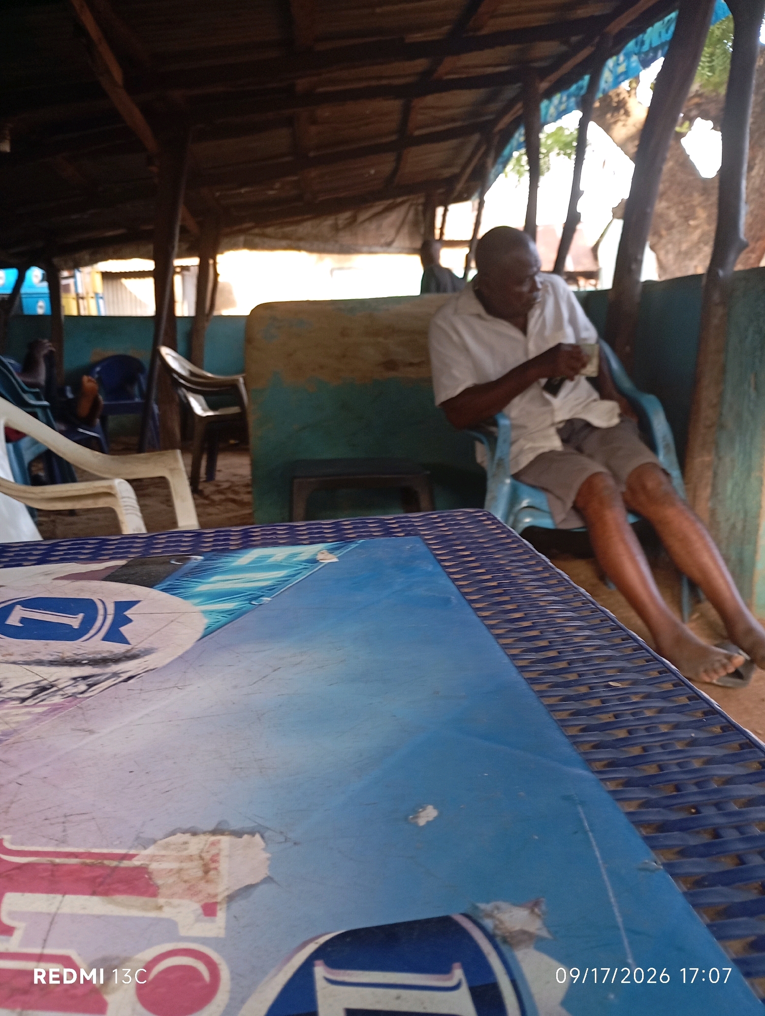 The image depicts an outdoor shaded seating area, likely a local eatery or bar, in Shendam, Nigeria. A middle-aged Black man, wearing a white short-sleeved shirt and gray shorts, is seated in a blue plastic chair, holding a cup. He appears to be looking down and to the right. The seating area is covered by a corrugated iron roof supported by wooden beams. In the foreground, a blue table with a patterned surface is visible. In the background, other chairs and tables, as well as other patrons, are dimly visible. The ground is dirt. The overall scene suggests a casual, everyday setting.