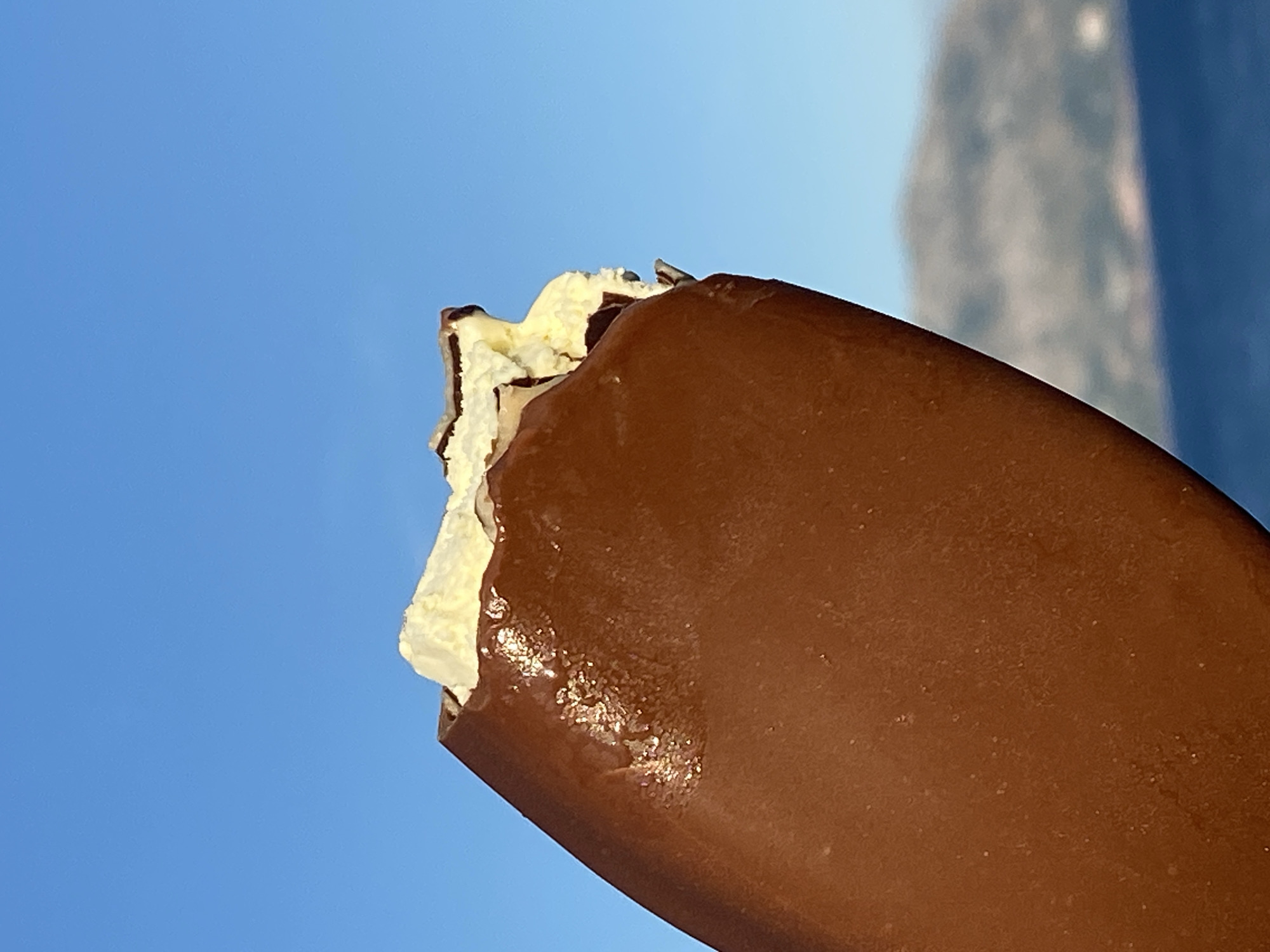 A close-up, eye-level shot focuses on a partially eaten chocolate-covered ice cream bar held against a bright blue sky. The ice cream bar, angled from the lower left corner upwards, is mostly coated in dark chocolate, which has a slightly glossy finish with some visible imperfections. The top of the bar reveals a bite taken out, exposing the creamy white ice cream and dark chocolate streaks within. The background is blurred, showing a hazy, distant landscape of mountains and a deep blue body of water, suggesting a coastal or seaside location. The scene does not contain any people or specific urban elements that would indicate Prijevor, Montenegro, or any other city.
