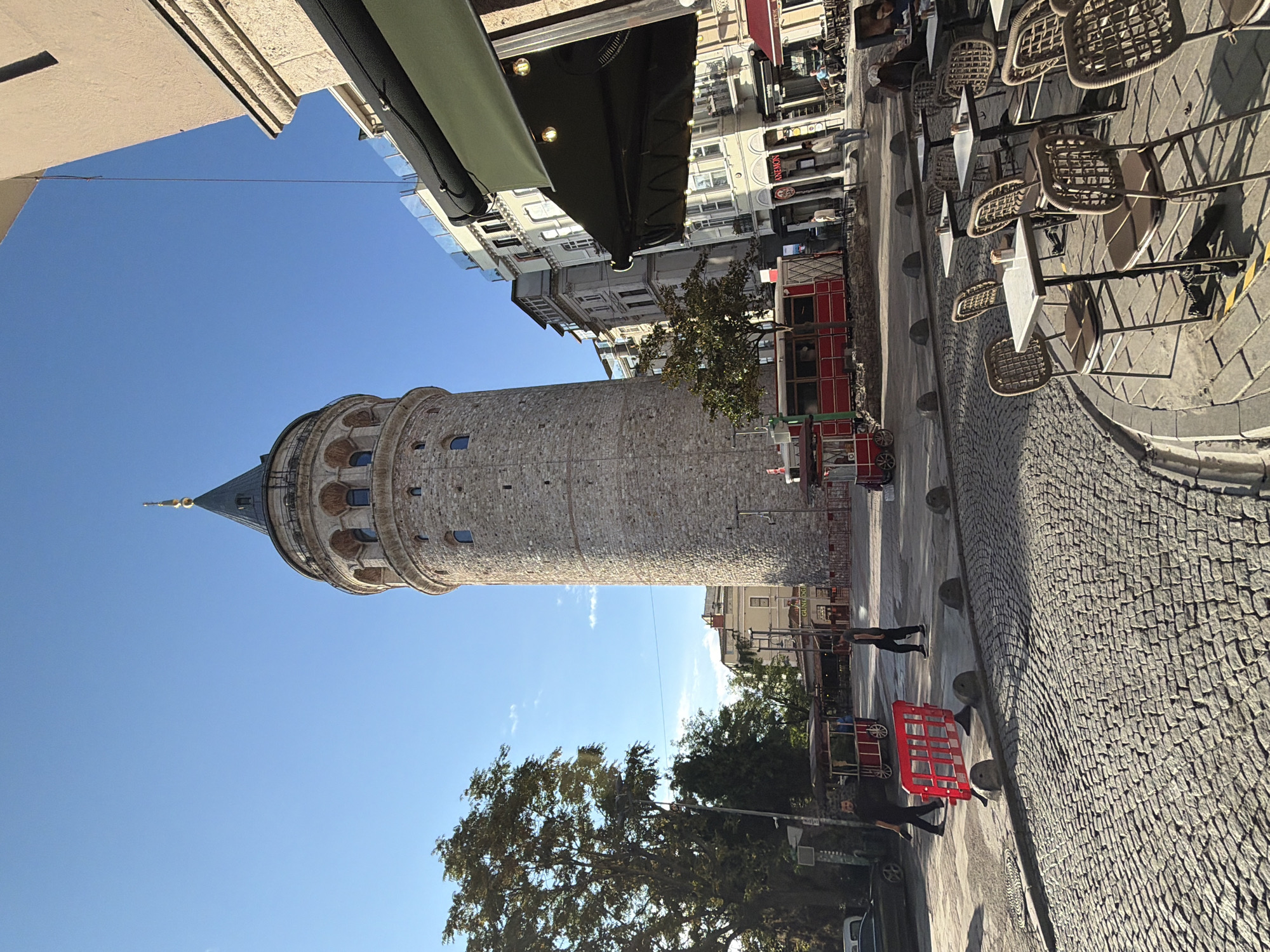 The image captures a view of the Galata Tower in Istanbul, Turkey, specifically in the Beyoğlu district. The tall, stone tower with its conical roof is the central focal point, standing against a clear blue sky. In the foreground, a cobblestone street curves towards the right, lined with outdoor cafe seating. Several tables and chairs, made of woven material and metal, are arranged on the sidewalk.
To the left of the tower, a large tree with dense foliage provides shade. In the middle ground, there are two red, trolley-like structures, possibly food or souvenir vendors, positioned on the street. Several people are visible walking in the area, including two men in the left mid-ground and a person walking towards the tower from the right mid-ground. Buildings with multiple stories, typical of urban architecture, line the street behind the tower and to the right. The atmosphere suggests a daytime scene with good weather.