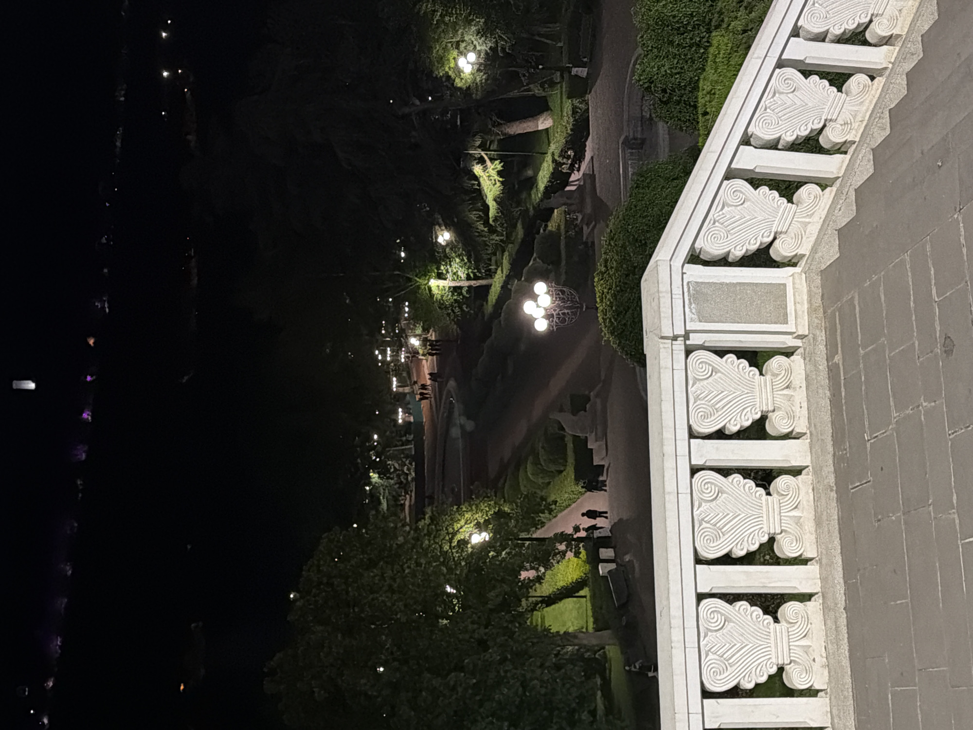 From an elevated perspective, the nighttime scene captures Vake Park in Tbilisi, Georgia. A decorative white stone balustrade extends down the right side of the image. Beyond the balustrade, a pathway curves through the park, illuminated by spherical streetlights. Several indistinct figures walk along the path. Trees and manicured shrubs line the path, contributing to the park's ambiance. In the background, a dark expanse suggests a lake or river, with faint lights reflecting on the water's surface. The overall atmosphere is peaceful.