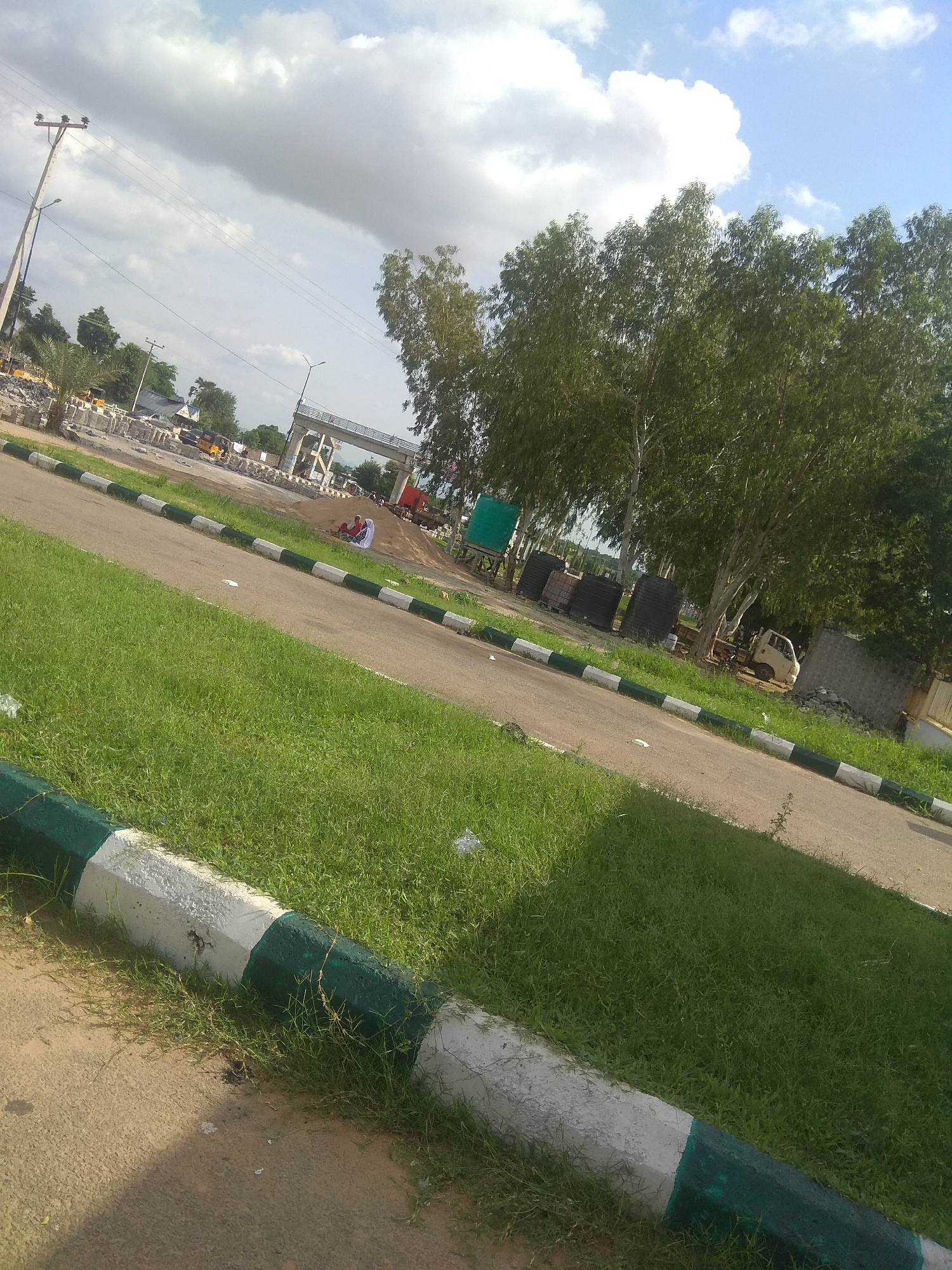 This is a photograph taken in Jalingo, Nigeria, likely from an elevated vantage point looking towards a road and surrounding construction or development. In the foreground, a grassy area borders a paved road. The road is marked with alternating green and white painted curbs. Further down the road, there are several people sitting by a construction site, with piles of building materials and some heavy machinery visible. A large, unfinished bridge or overpass structure is present in the middle ground, and trees line the background. The sky is partly cloudy with a mix of blue sky and white clouds. Electrical poles and wires are also visible throughout the scene.