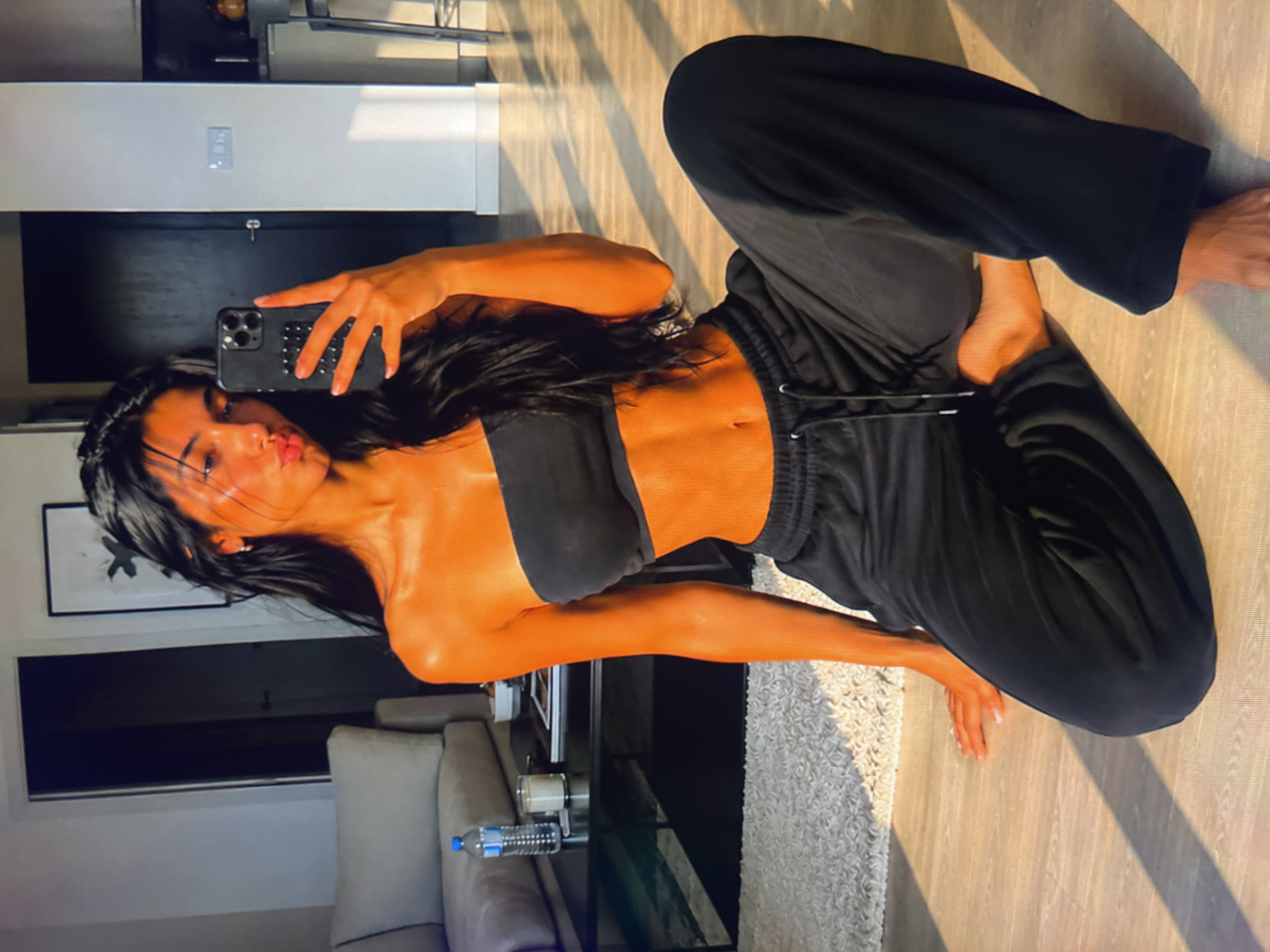 A woman with long, dark hair is kneeling on a wooden floor, holding a smartphone to take a selfie. She is wearing a black strapless top and black sweatpants. The room appears to be a living room with a sofa, coffee table, and a rug visible in the background. Sunlight streams in from the right, casting long shadows across the floor.