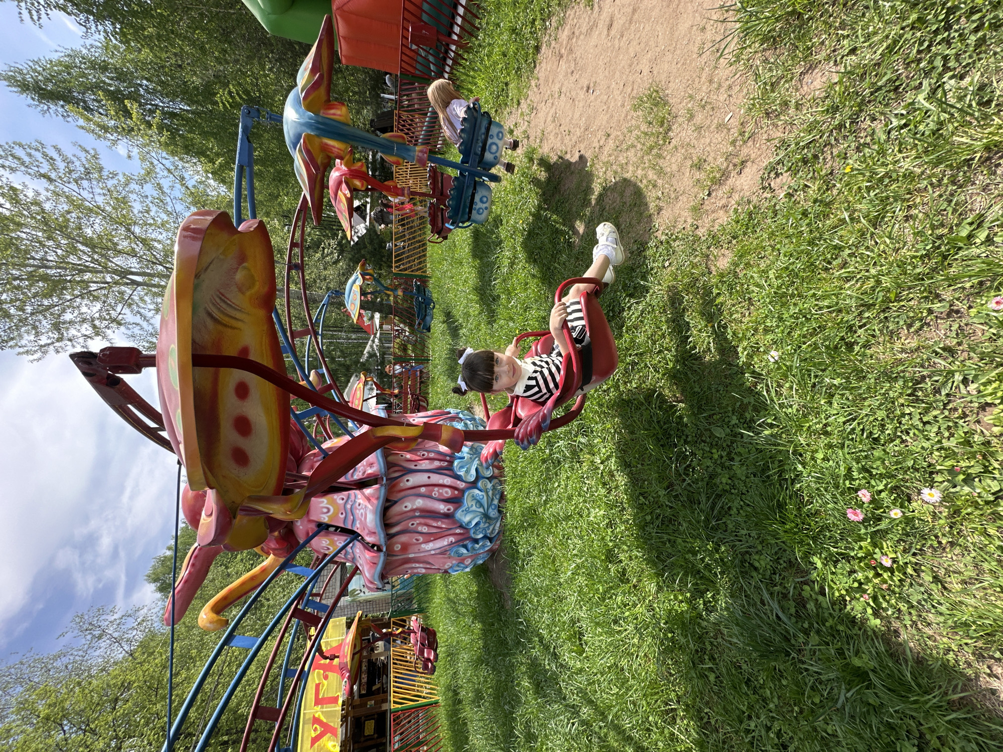 A child is riding a carousel at the Park of Culture and Rest in the city of Chaikovsky, Russia. The child is wearing a striped shirt and white shoes and is seated in a red seat attached to the carousel. The carousel is colorful with various shapes and figures. The ground is covered in grass and there are trees in the background. Other children can be seen riding other parts of the carousel in the background.