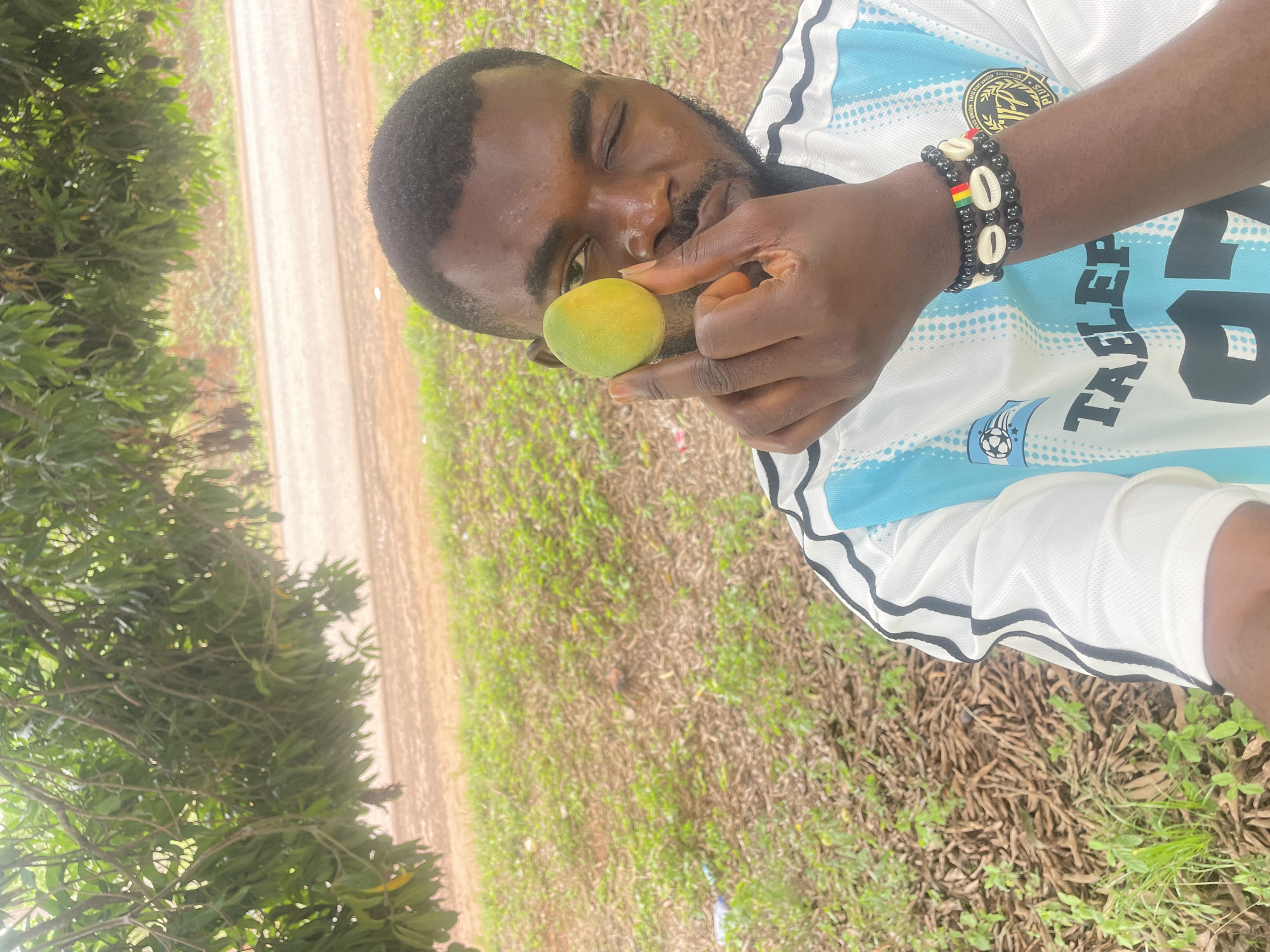 An African man stands in the Ogbe, Nigeria, countryside, holding a green mango up to his face. He wears a white sports jersey with blue and black trim. Two beaded bracelets are on his left wrist. He stands beside a dirt road bordered by greenery.