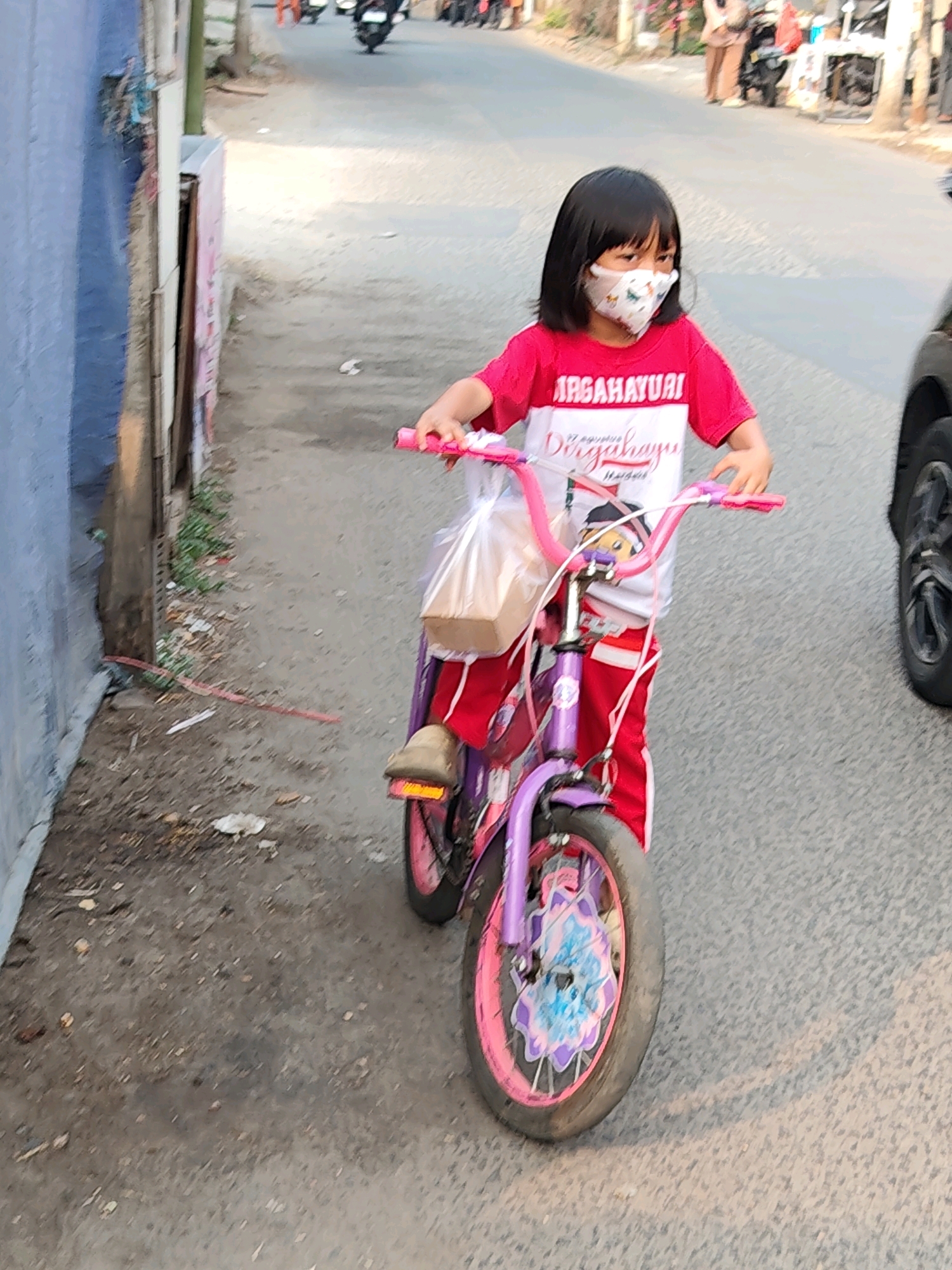 A young girl wearing a face mask and a red and white shirt is riding a purple bicycle on a paved street in Bekasi, Indonesia. The girl has black hair and is holding the handlebars of the bicycle. A plastic bag containing a light brown object is attached to the handlebars. The bicycle has pink wheels and a decorative element on the front wheel. The street is lined with a blue wall on the left and appears to be an urban area with some debris on the ground. Other people and vehicles are visible in the background, suggesting a public street.