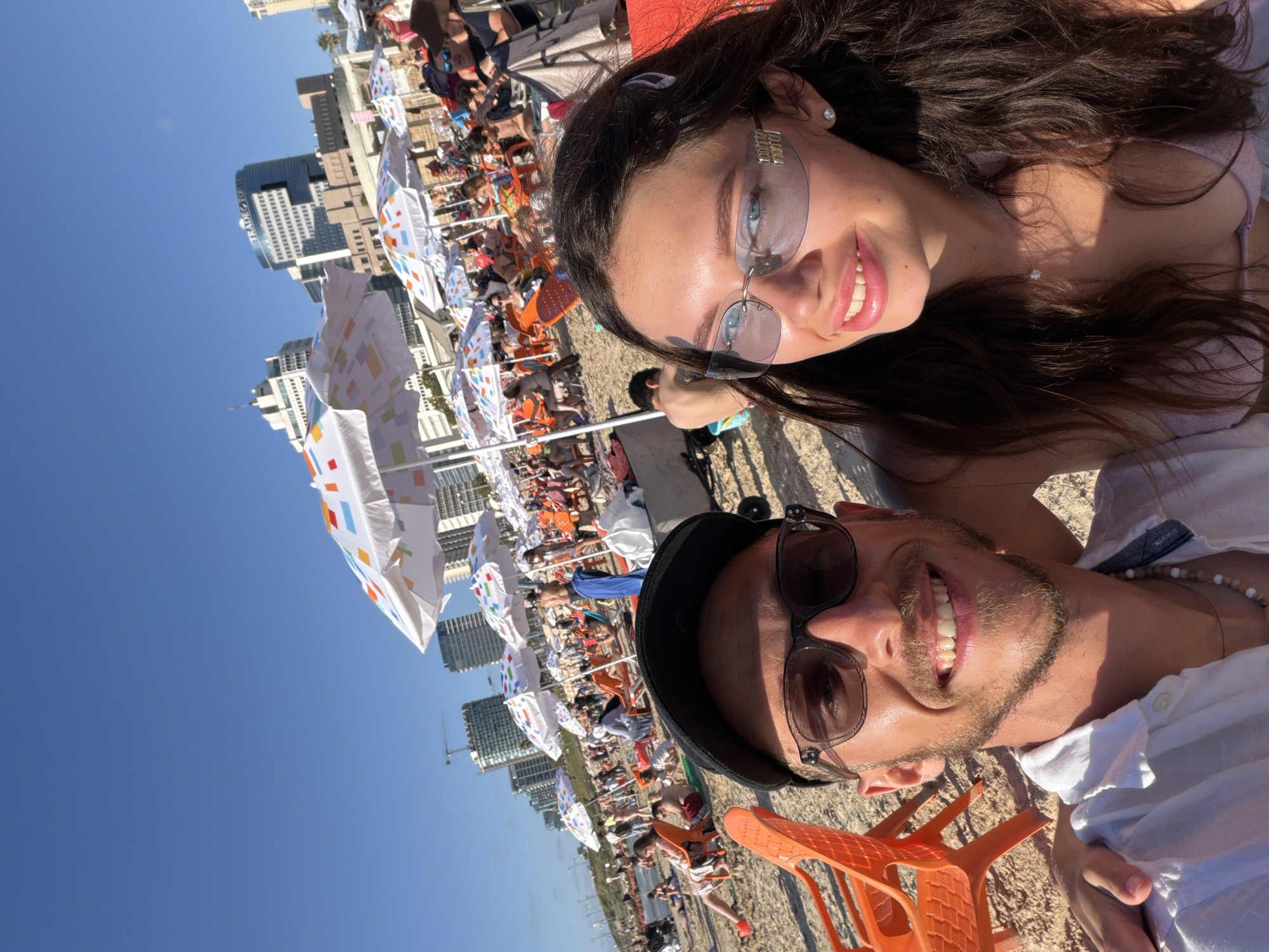 This is a selfie taken on a crowded beach in Tel-Aviv, Israel. The foreground features two young adults, a man and a woman, smiling at the camera. The man, wearing a white shirt and a black cap, is on the left, and the woman, wearing a light pink bikini top and sunglasses, is on the right. Behind them, the beach is filled with people relaxing under numerous colorful umbrellas, with the cityscape of Tel-Aviv visible in the background, including tall buildings and a construction crane. The sky is clear and blue, indicating a sunny day. The scene captures a lively atmosphere of a popular beach destination.