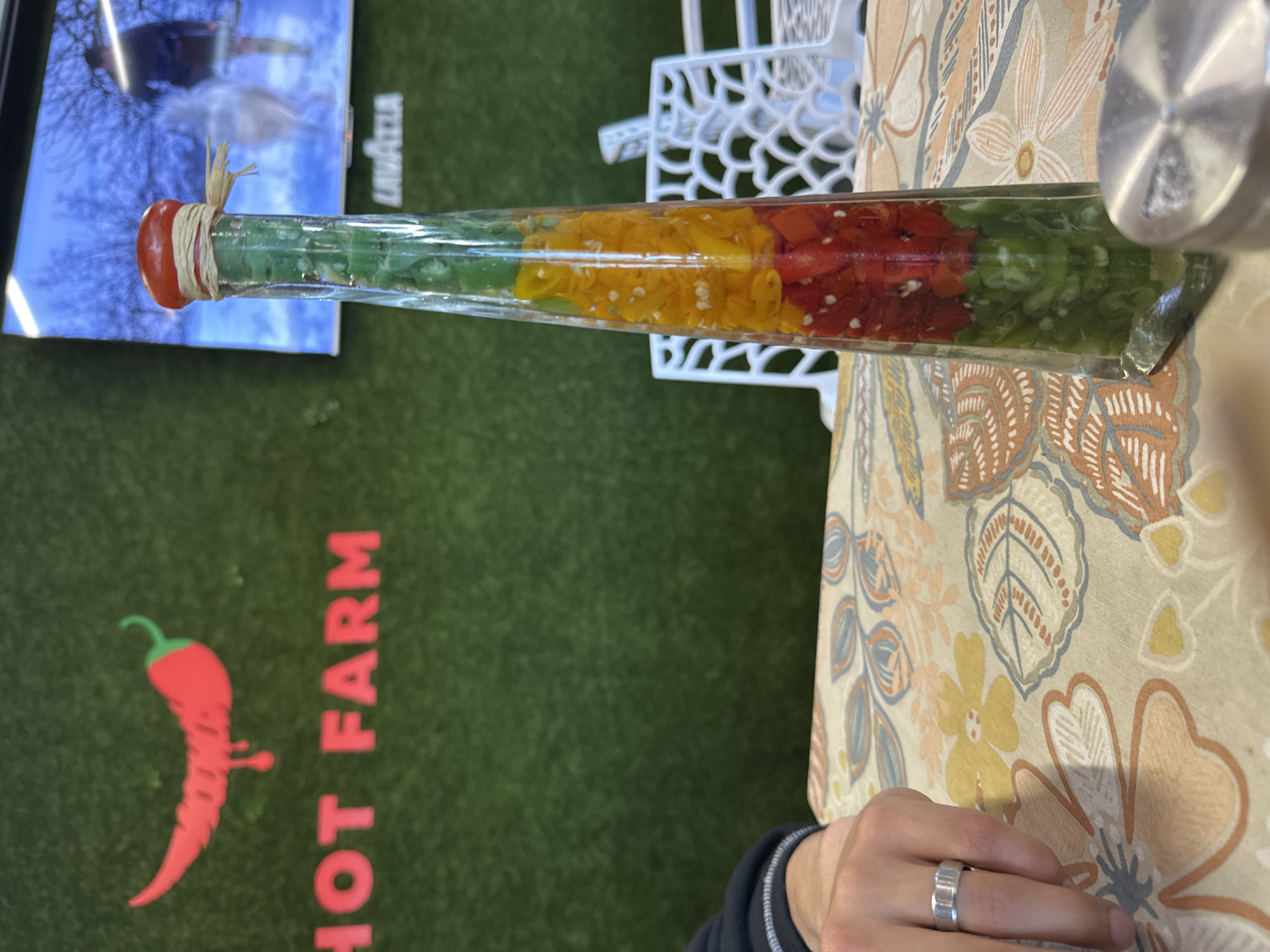 The image shows a long, slender glass bottle filled with layers of green, yellow, and red chopped peppers, sealed with a red cap and tied with twine, resting on a table covered with a floral-patterned tablecloth. A white decorative chair back is visible near the bottle. A sign for "Hot Farm" with a chili pepper logo is visible behind the table. In the background, there is a television screen and a person wearing a ring and a black jacket sleeve. The image was taken in Beloslav, Bulgaria.