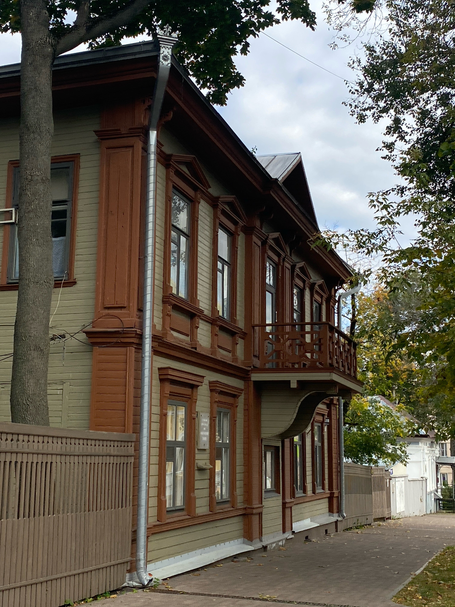 An external, eye-level, medium shot captures the side of a two-story wooden house with green siding and brown trim, situated in Nizhny Novgorod, Russia. The house features multiple windows with brown frames, some with decorative cornices, and a second-story balcony with an ornate wooden railing. A large tree trunk with rough bark stands to the left of the house, and a tall, silver drainpipe runs vertically down the side of the building. A wooden fence with vertical slats borders the property on the left, and a paved walkway extends along the right side of the house. Autumn leaves are scattered on the ground. The sky above is overcast. The scene appears quiet and undisturbed, with no people or animals visible.