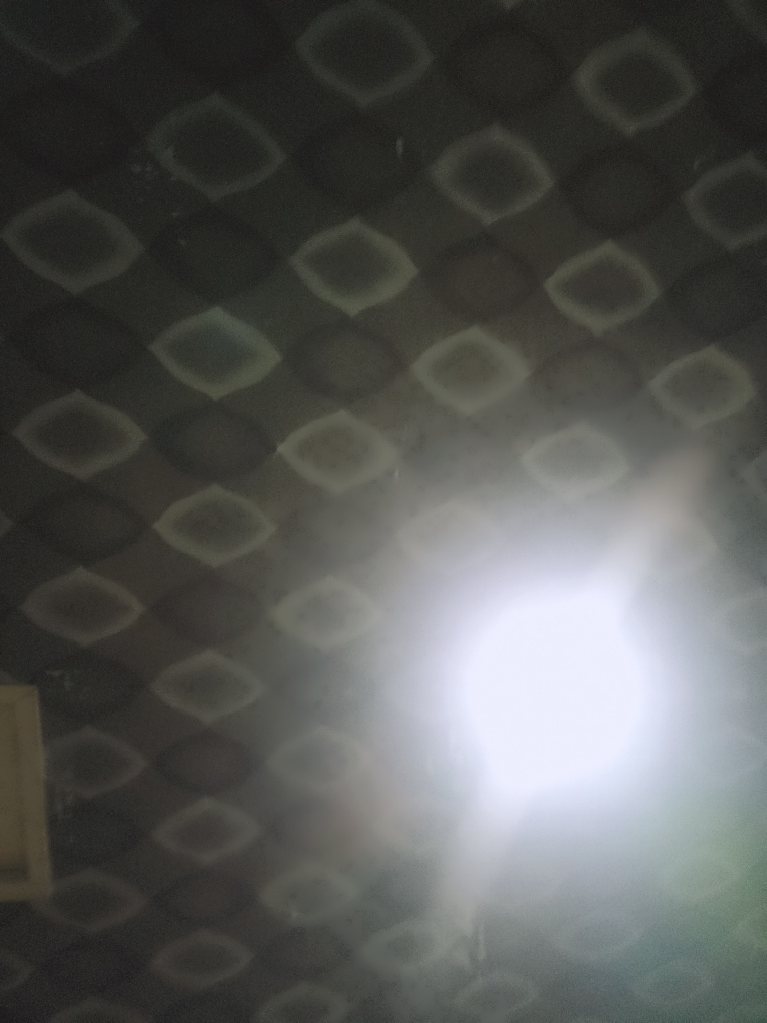 The image is a close-up, low-light photograph of a textured dark surface with a repeating diamond pattern. A bright light source, possibly a flash or direct light, is positioned in the lower right quadrant, causing significant overexposure and glare. This bright light washes out the details of the texture in its vicinity, creating a hazy effect. In the lower left corner, a small portion of a pale, rectangular object is visible, possibly a wall or part of furniture, partially obscured by the glare. The overall impression is abstract, with the strong light overpowering the subtle pattern of the surface. The location is Unknown, Unknown.