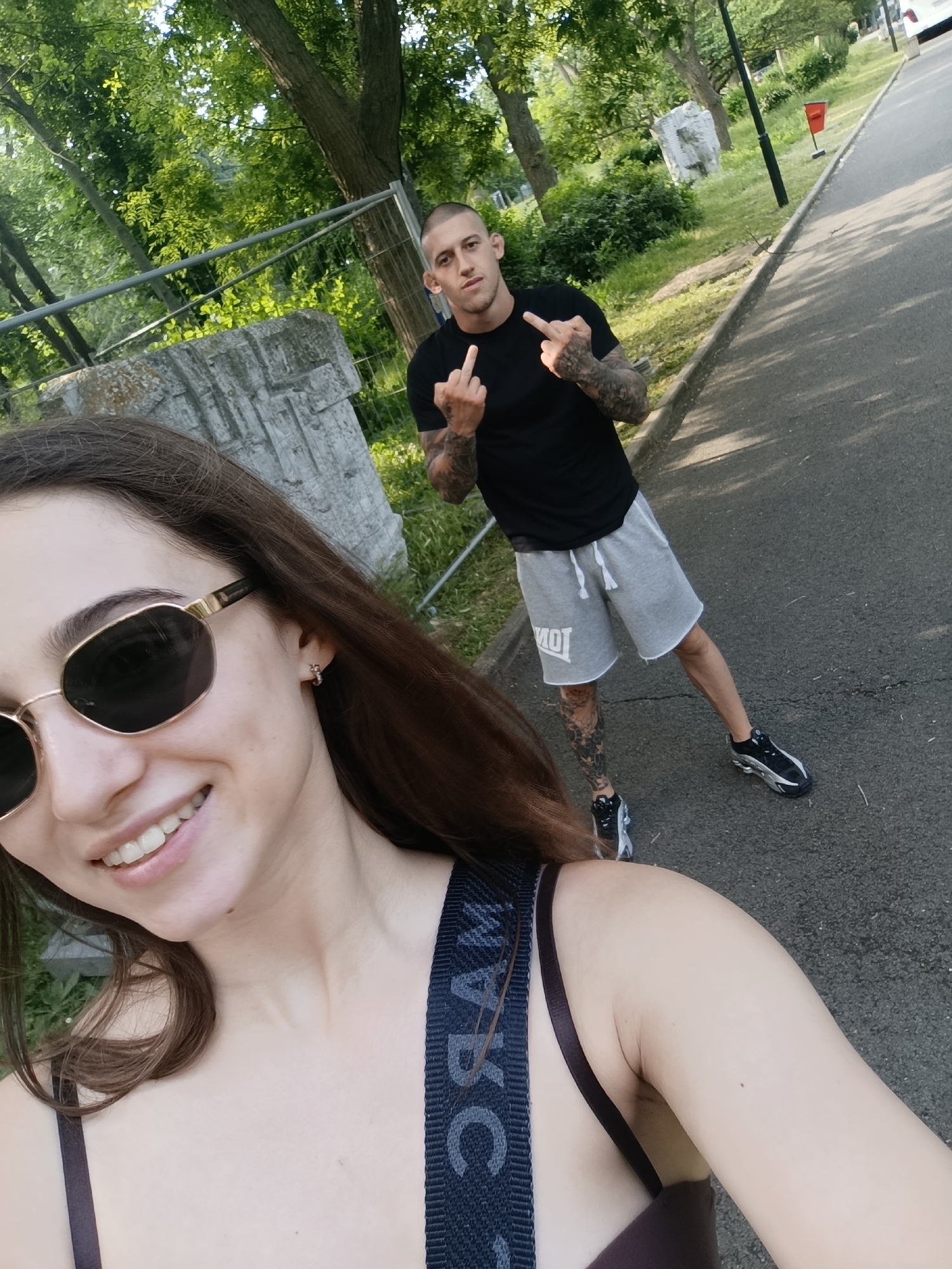 The image captures two individuals outdoors on a paved path. In the immediate foreground, a female with long brown hair, wearing gold-framed sunglasses and a dark top with a visible "MARC" branded strap, is smiling. A small hoop earring is visible in her right ear. She appears to be taking a selfie, holding the camera towards herself.
In the mid-ground, positioned to the right and behind the female, a male with short hair stands on the paved surface. He is wearing a black t-shirt, grey shorts with a "MOT" logo on the left leg, and black and silver athletic shoes. Numerous tattoos are visible on both his arms and legs. He is captured with both hands raised, each extending the middle finger towards the camera.
The environment features a paved road or pathway bordered by green grass and numerous trees with dense foliage. A metal fence runs horizontally across the mid-ground, behind which a large, light-colored stone structure or monument with carvings is partially visible. Further in the background, a black lamppost and a red trash can are present alongside the road, with a white vehicle partially visible in the upper right corner. The scene is illuminated by natural daylight. The specific location within the city of Burgas, Bulgaria, cannot be identified from the visual information provided.