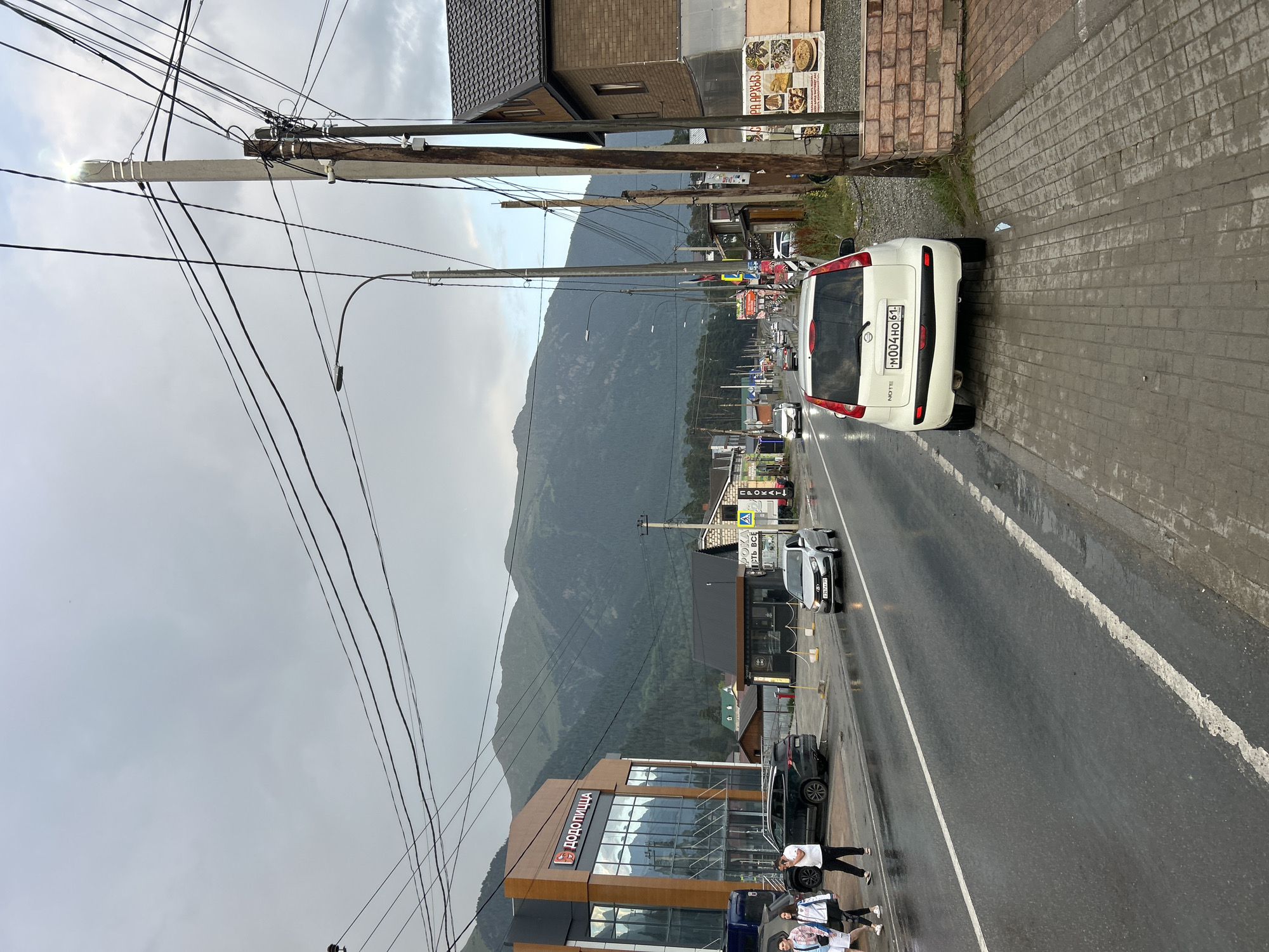 A street view in Arkhyz, Russia, features a wet asphalt road lined with buildings and a backdrop of imposing mountains. On the left, a modern building with large glass windows displays the logo "ДОДОПИЦЦА" (Dodo Pizza). Several people are walking on the sidewalk in front of this building. To the right of the road, a white Nissan Note with license plate "M004HO 61" is parked. Further down the street, other vehicles are present, and power lines are strung across poles. The sky is overcast.