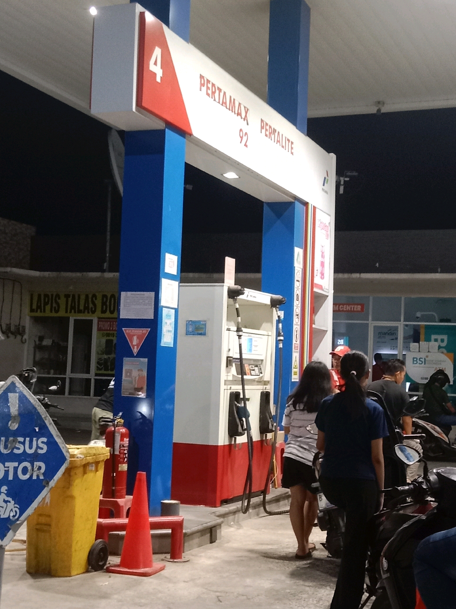 This image captures a nighttime scene at a Pertamina gas station in South Tangerang, Indonesia. A gas pump with "PERTAMAX PERTALITE 92" signage is prominent, with a blue and white structure housing the pump. Several people are present, some on motorcycles, suggesting a refueling or waiting activity. A yellow trash bin and a red traffic cone are visible near the pump, along with a fire extinguisher. In the background, a building with "M CENTER" and "mandiri" signage indicates a commercial area. The ground is paved, and the overall lighting suggests artificial illumination typical of a gas station at night.