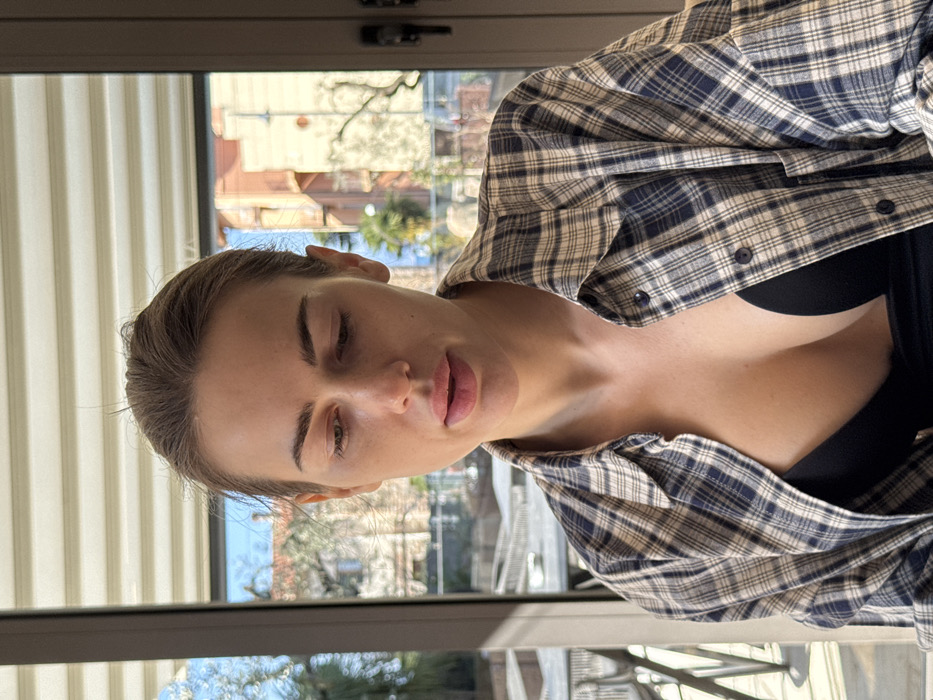 A female individual is centrally framed from the upper chest, seated indoors. She has light brown hair pulled back from her face, with eyes cast downwards and to her left. Her lips are slightly parted, revealing no apparent cosmetic enhancements or makeup on her face. She wears an unbuttoned blue and beige plaid collared shirt over a black undergarment, which appears to be a bra or deep-cut top. A black adhesive breast support strip is visible on her left breast. Behind the individual, a large window provides a view of an outdoor environment. The background, largely out of focus, reveals clear blue skies, indicative of a sunny day. Residential buildings with terracotta-colored facades and roofs are visible, along with green foliage including palm trees and bare deciduous trees. A body of water, consistent with a large lake, is visible in the mid-distance. Outdoor tables and chairs are partially visible on what appears to be a terrace or balcony below. The scene is located in Albisano, Italy.