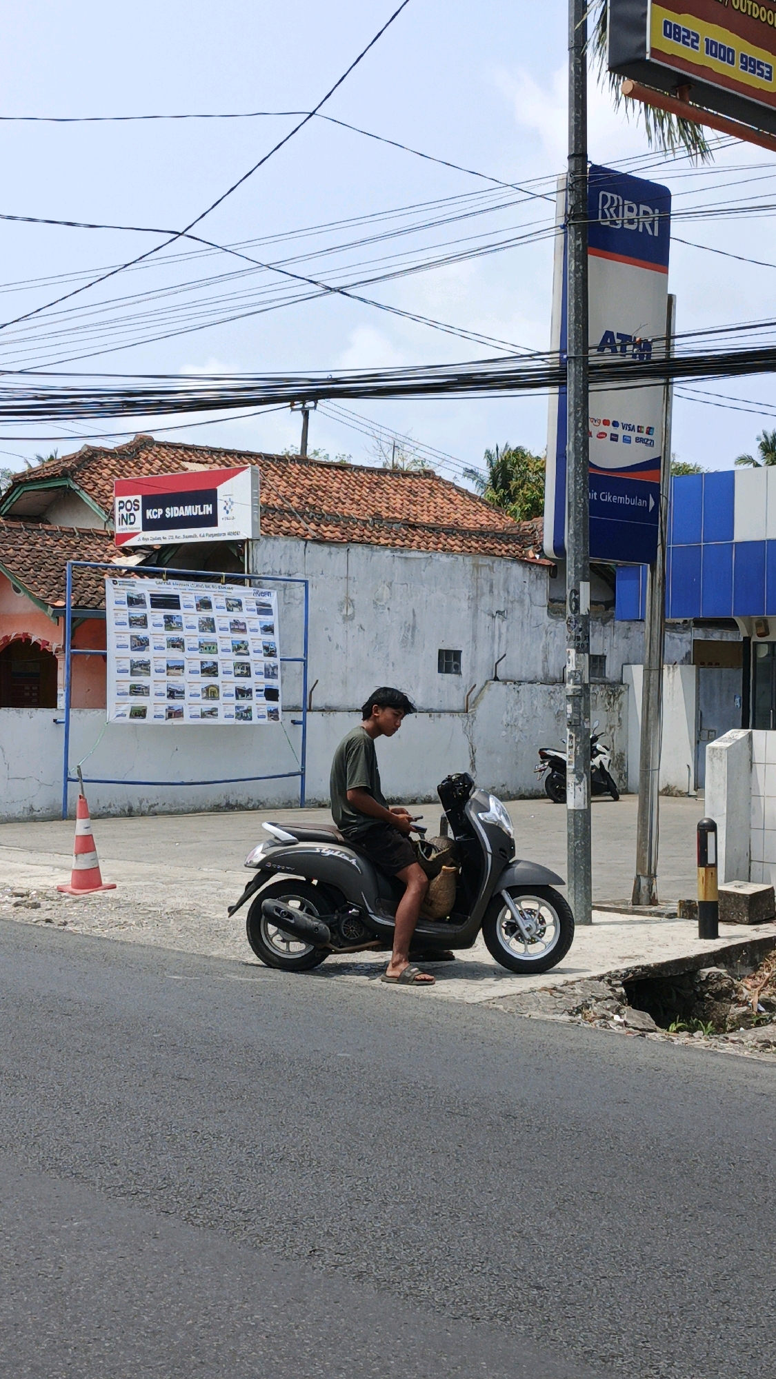 The image shows a young man sitting on a dark gray scooter parked on the side of a road in Desa Wonoharjo, Indonesia. He is wearing a dark green t-shirt and dark shorts, and appears to be looking at his phone. The scooter has a wicker basket attached to the front. Behind the scooter, a large poster displaying multiple small photographs of houses is mounted on a blue metal frame. To the left of the poster is a red and white traffic cone. Further to the left, a building with a red tiled roof and white walls is visible, with a sign that reads "POS IND KCP SIDAMULIH". To the right of the scooter, a tall blue sign with the BRI logo and "ATM" is displayed. The background includes overhead electrical wires, a blue building, and trees. The ground is paved with asphalt and concrete.