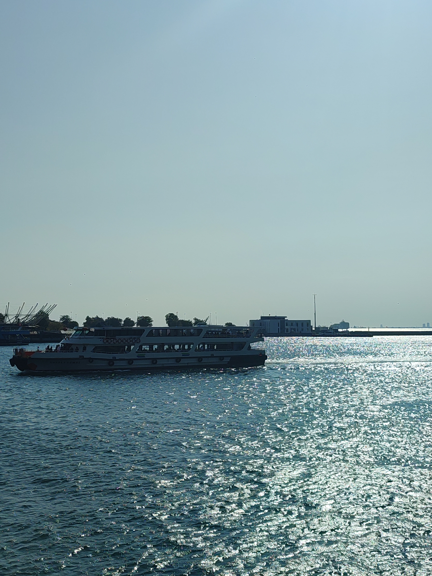A wide, horizontal shot captures a passenger ferry sailing across a shimmering, sunlit sea. The ferry, a long, white vessel with multiple decks and rows of windows, is the central subject, positioned on the left side of the frame and moving from left to right. Several people are visible on the ferry's decks. In the background, beyond the ferry and the water, a shoreline is visible with trees and buildings. A long pier extends into the water from the right side of the frame, and on it, there is a modern, multi-story building with a tall pole. Further in the distance, more buildings and structures can be seen along the horizon, hinting at a city skyline. The sky is a pale, clear blue, devoid of clouds. The water reflects the sunlight, creating a bright, sparkling surface. To the left of the ferry, some older maritime structures, possibly cranes or masts, are visible, suggesting a harbor area. The scene appears to be a sunny day, likely in the city of Kadıköy, Turkey, given the characteristic ferry transportation and the urban shoreline.