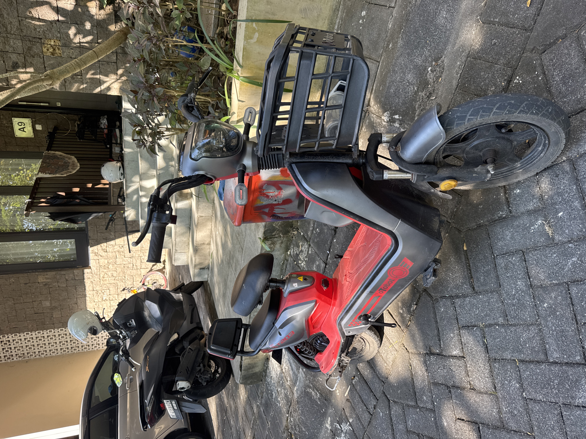 The image depicts an outdoor scene in Kuta Selatan, Indonesia, under bright daylight, featuring multiple vehicles and a residential building entrance. In the foreground, a red and grey electric scooter is parked on grey interlocking pavers, angled slightly to the left. The scooter features a black front basket containing a clear plastic bottle, a clear headlight, black handlebars, and "Tobot" branding visible on its red footboard. A red container with graffiti, possibly reading "RED," is positioned between the handlebars and the seat. The seat is black with a small backrest.
Behind the electric scooter, a black conventional scooter, identified as a Honda PCX by its visible lettering and design, is parked. A white helmet rests on its handlebars. Its license plate, "DK 4168 FCN," confirms its origin from Bali, Indonesia. A silver four-door car is partially visible behind the black scooter. A red bicycle is discernible further back, leaning against a wall near the building.
The background features a building with a stone-clad exterior. A set of concrete steps leads to a dark brown door marked with a white sign displaying "A9." Another white helmet is positioned on the steps. A shoe rack with footwear is visible next to the door. A railing near the steps holds an umbrella and what appears to be a small rug. Green and reddish-purple foliage grows in garden beds along the path and near the building. A tree trunk is present on the right, casting distinct shadows across the scene. The ground is paved with interlocking grey and dark grey rectangular blocks in a herringbone pattern.