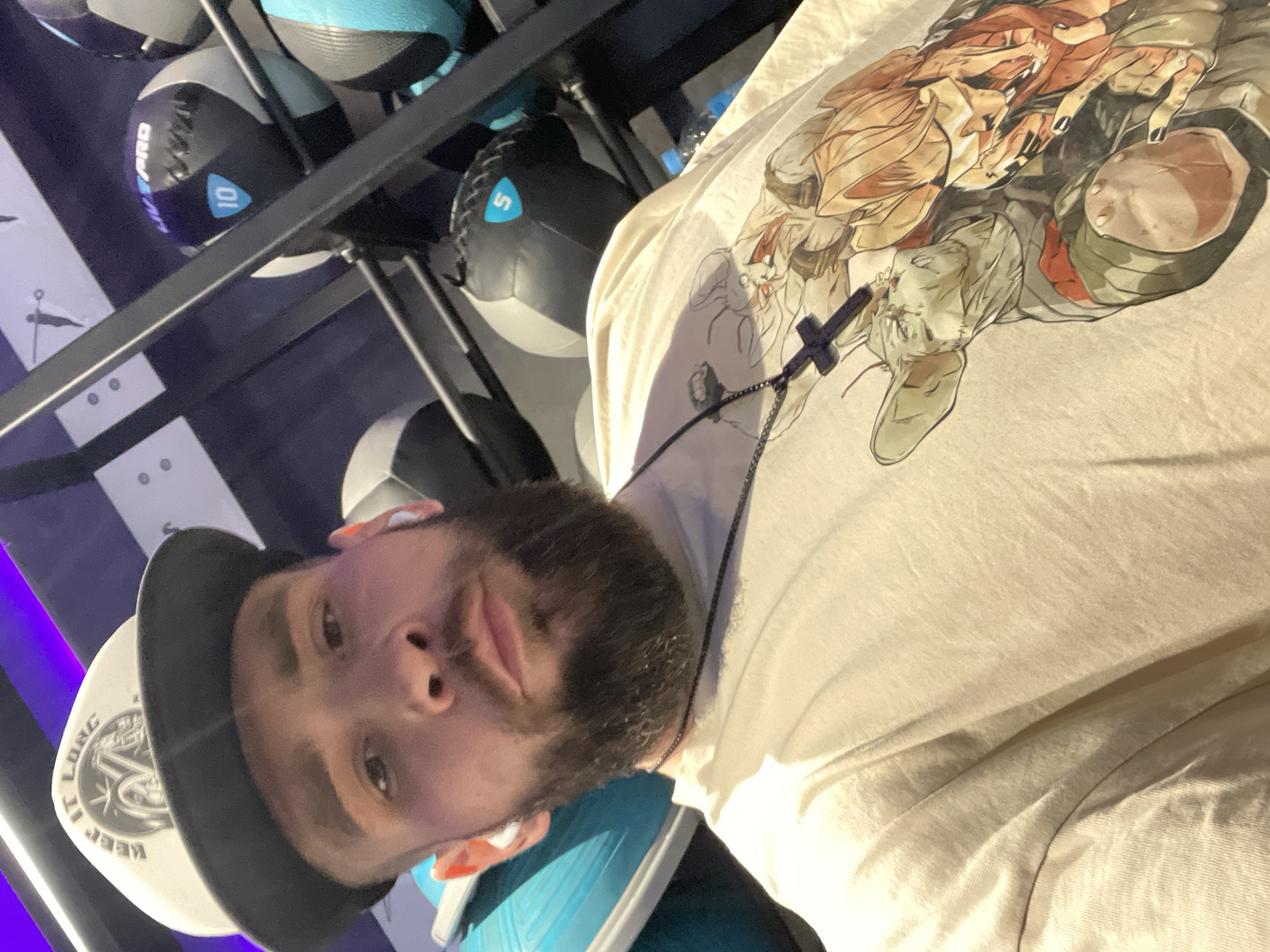 A man is taking a selfie in a gym. He is wearing a white baseball cap with black lettering that says "KEEP IT LONG" and a light-colored t-shirt with a graphic print of characters. He has a beard and is wearing white wireless earbuds. In the background, there are shelves filled with various medicine balls, some with numbers and branding visible. The lighting in the gym appears to be a mix of fluorescent and possibly some colored accent lights. There are no specific details in the image to suggest it is located in Khabarovsk, Russia.