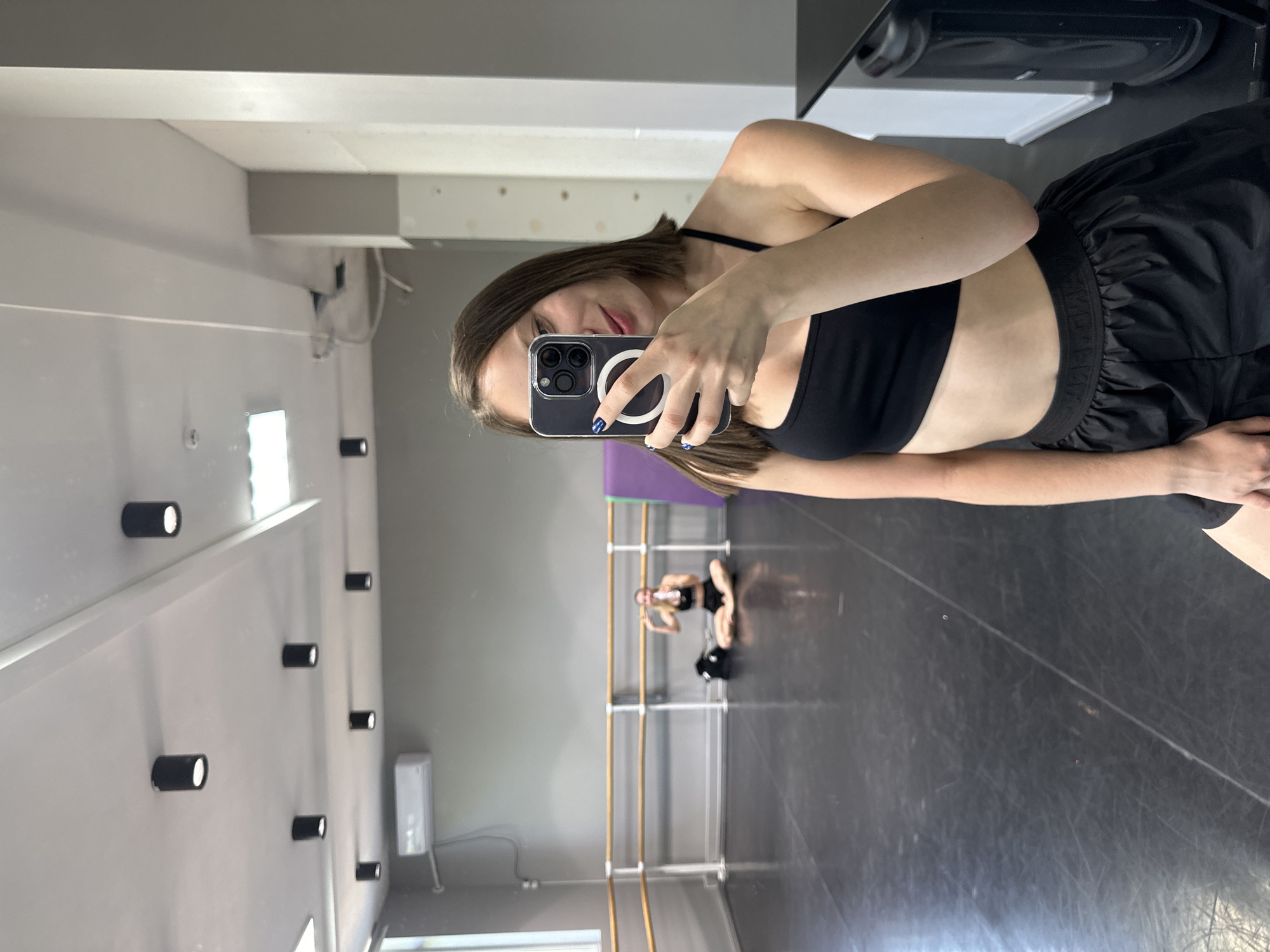 A young woman is taking a mirror selfie in what appears to be a dance studio. She is wearing a black sports bra and black shorts, with her hair down. In the background, another person is visible sitting on the floor in a similar pose. The studio has wooden barre railings along the walls and a large mirror reflecting the scene. There are also several cylindrical lights mounted on the wall. The overall lighting suggests it is daytime. The woman's expression is neutral as she captures the photo. The location is identified as Kryvyi Rih, Ukraine.