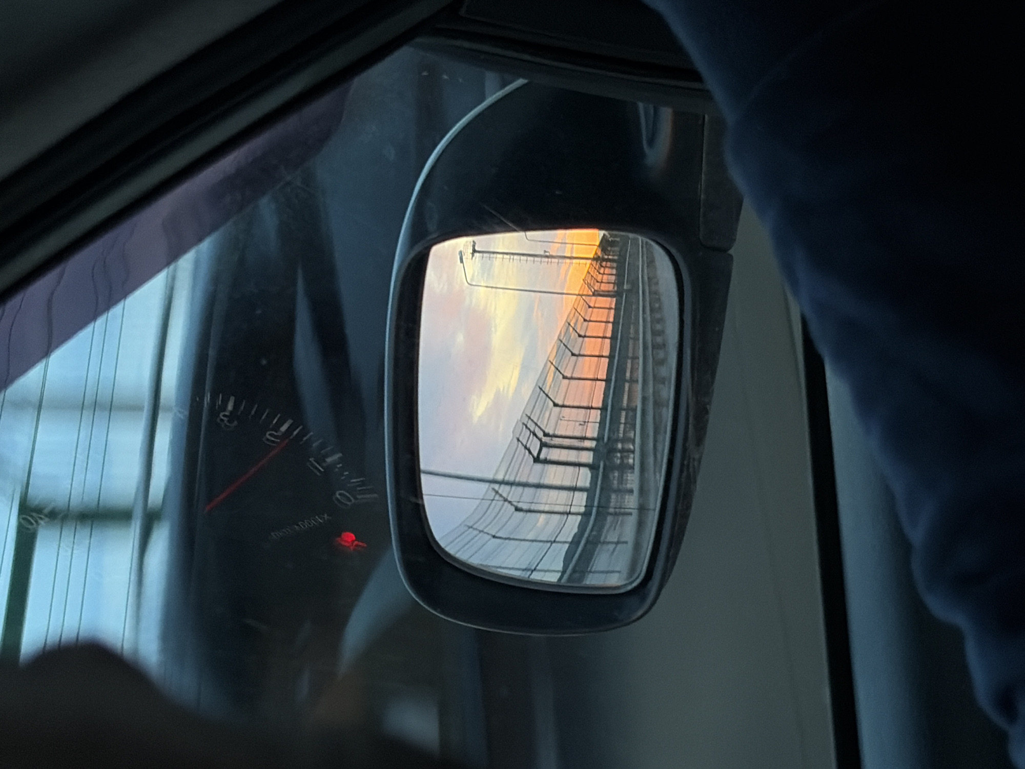 From inside a vehicle, a side mirror reflects a bridge at sunset. The bridge has a pedestrian walkway with a railing, and streetlights are visible. The sky is orange and pink, indicating sunset or sunrise. Inside the car, a dashboard gauge is partially visible, with a red indicator light illuminated. A blue sleeve is also in view, suggesting a person is present in the vehicle. The exact location of the bridge within Chaikovsky, Russia, is not discernible from the provided image.