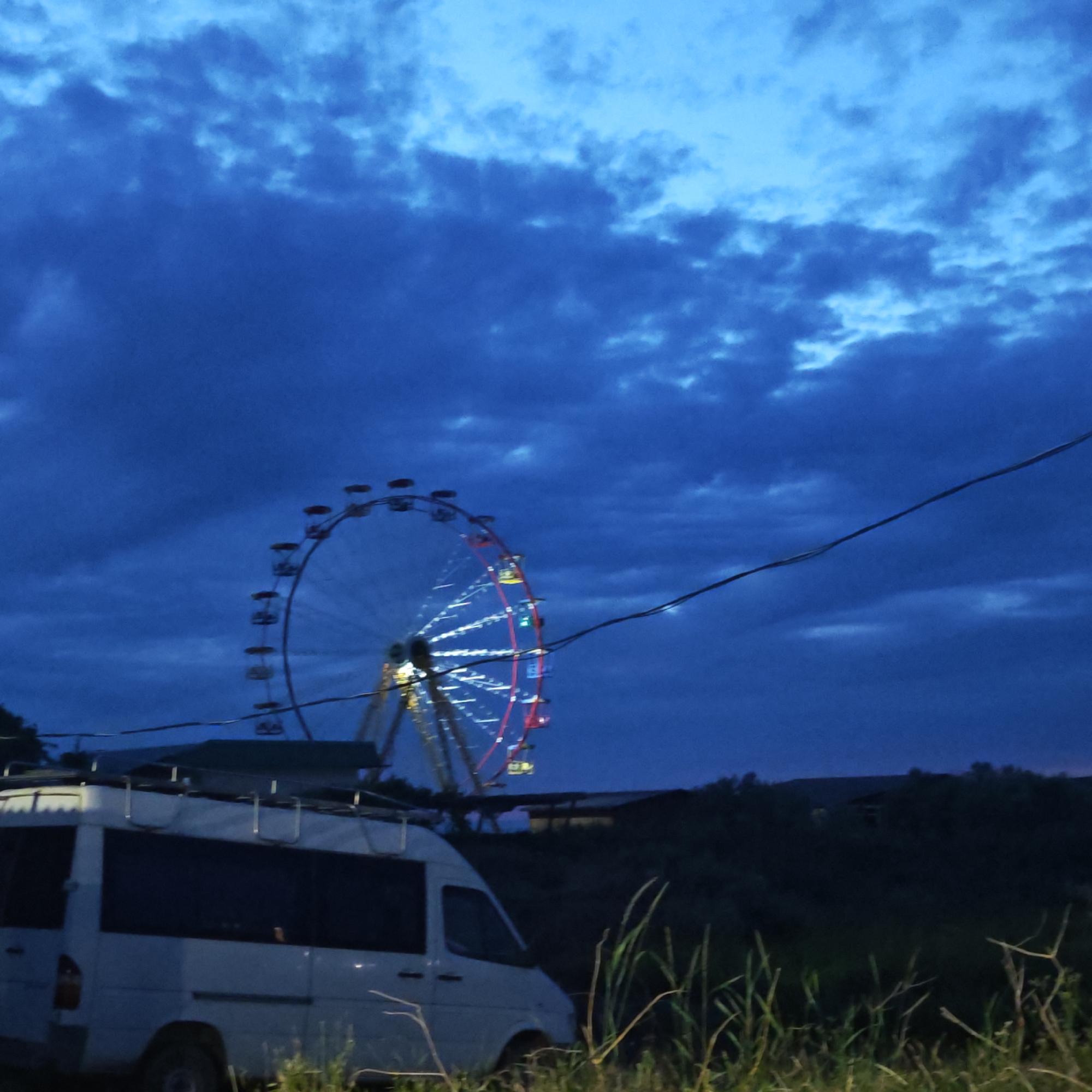 The image captures a nocturnal or late dusk scene, dominated by a large, illuminated Ferris wheel against a dark, cloudy blue sky. The Ferris wheel is partially visible, with its structure lit by numerous white and red lights, suggesting it is either operational or recently lit up in an amusement park or fairground setting.
In the foreground, a white van, possibly a camper or utility vehicle, is parked on the left side, featuring a visible roof rack. Tall, dark green grass occupies the bottom edge of the frame, indicating natural or unkempt surroundings. A dark, possibly electrical, cable stretches diagonally across the upper middle of the sky, passing in front of the Ferris wheel.
Beyond the van and Ferris wheel, the background consists of dark, silhouetted hills or dense bushes under the deep blue, cloud-covered sky. The artificial lights of the Ferris wheel contrast sharply with the prevailing darkness of the environment. No people are visible, and there is no discernible text in the image. The mood is serene and somewhat atmospheric, characteristic of twilight or nighttime. The scene is set in what appears to be a semi-rural or recreational area, consistent with the provided location context of городское поселение Щелкино, Ukraine.