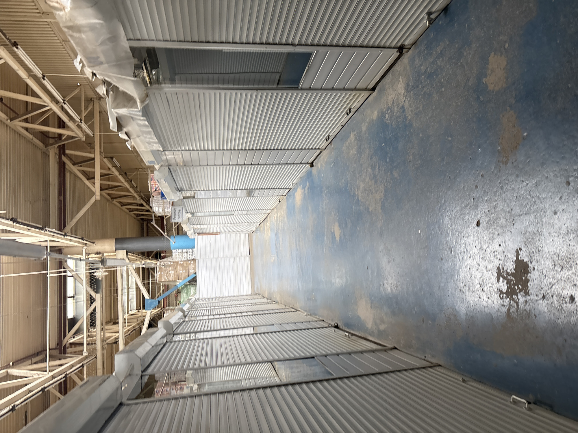 The image shows a long, indoor market hallway in Pastavy, Belarus. On either side of the hallway are rows of closed stalls with metal roll-up doors. The floor is a worn blue surface with some dark stains and dusty patches. The ceiling is a high industrial structure with exposed beams, pipes, and lighting. In the background, there are some stacks of boxes and what appear to be white partition walls. The overall impression is of an empty or closed market.