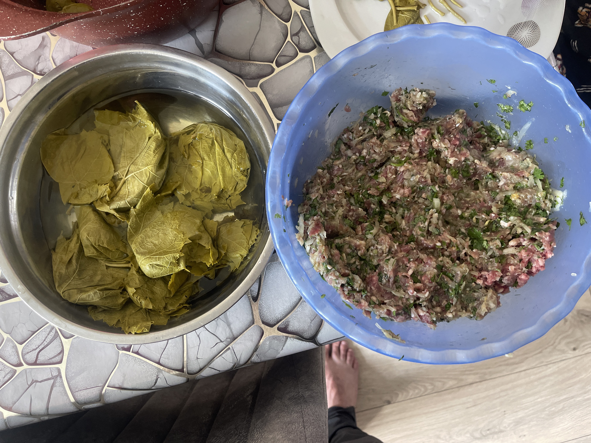 A high-angle medium shot shows a table setup with ingredients for dolma, a traditional Azerbaijani dish. In the upper portion of the image, a silver bowl contains blanched grape leaves submerged in water. Adjacent to it, a partially visible reddish-brown pot also holds grape leaves. The lower portion of the image features a blue plastic bowl filled with the dolma filling, a mixture of ground meat, rice, herbs, and seasonings. A small white plate with some yellow, possibly pickled, items is visible to the right of the blue bowl. A person's bare foot is visible on the wooden floor in the lower left corner, suggesting the preparation is taking place in a home environment, likely in Novxanı, Azerbaijan, given the context. The table surface is covered with a patterned tablecloth.
