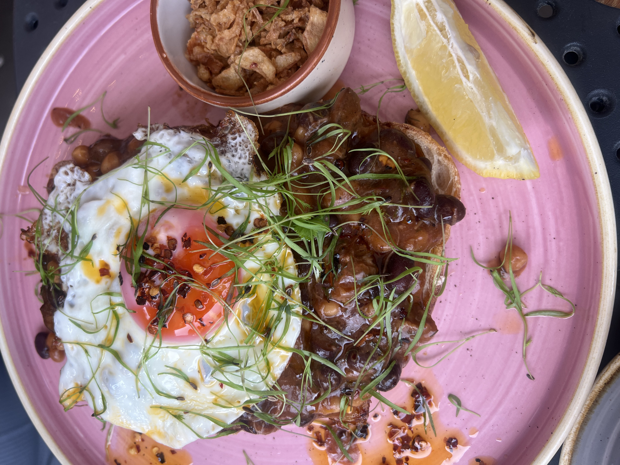 Here is a detailed description of the image:
The image is a close-up, overhead shot of a meal presented on a pink plate with a textured, slightly uneven surface. The main focus is the food arrangement: A piece of toast is topped with a dark, possibly bean-based stew, a fried egg with a bright orange yolk, and scattered microgreens. On the side, there is a small bowl containing crispy fried onions. A slice of lemon sits next to the bowl of onions. There are some red pepper flakes on the egg and around the dish. The plate shows signs of spillage, with some sauce and greens scattered around the edge.
There are no people in the image. The scene appears to be a restaurant setting, although the wider environment is not visible. No further details about the specific location can be inferred from the image.