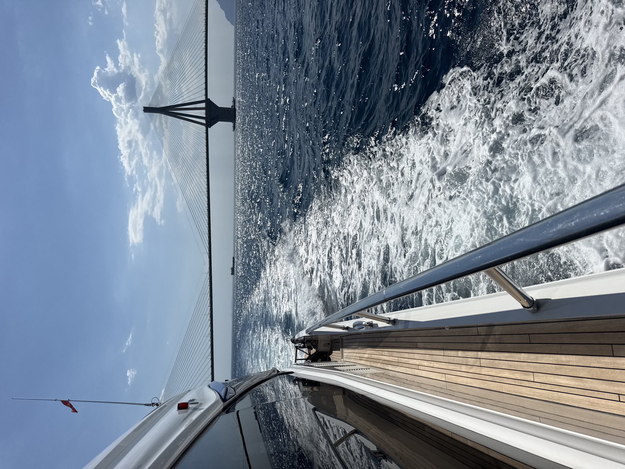 The image shows a view from a moving yacht, with the Municipal Unit of Rio, Greece in the background. The subjects include the yacht itself, the sea, and a large cable-stayed bridge. The scene is set on a bright, sunny day, with a clear blue sky and fluffy white clouds. The sea is dark blue and choppy, with white foam created by the yacht's movement.
The activity taking place is a boat ride or cruise, as indicated by the wake behind the yacht. The bridge, which is a prominent feature in the background, appears to be the Rio-Antirrio Bridge, a major landmark in the region. The yacht has a wooden deck and a metal railing, suggesting it is a luxury vessel. There are no people clearly visible, but the presence of a small red flag on an antenna suggests the yacht is in operation.
The time of day appears to be morning or afternoon, given the bright sunlight and clear sky. The weather is fair and pleasant, ideal for a boat trip. The location is clearly coastal, with the bridge spanning a body of water, likely the Gulf of Corinth. The overall mood is one of leisure and enjoyment of the scenic surroundings. No visible text is present in the image.