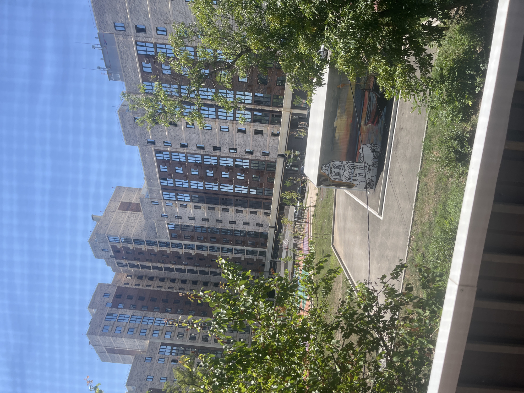 This is a medium shot photograph taken from a balcony, looking out at a modern urban residential area on a sunny day. In the background, a large, multi-story apartment complex with a beige brick facade and numerous windows dominates the scene. The building has a complex, stepped design with several protruding sections and variations in facade color, featuring darker brick accents. The sky is clear and bright blue, with a faint grid pattern visible across the image, suggesting it was taken through a screen or netting.
In the middle ground, a paved area, possibly a courtyard or parking lot, is visible. On this paved surface, there is a long, white, rectangular structure adorned with a mural. The mural depicts a sunset scene with a lighthouse or a church-like building on the left, and two boats on the water in the center. The mural is stylized, with a combination of outline drawing and colored elements. To the left of this structure, a colorful playground with geometric patterns is partially visible behind a fence.
The foreground and mid-ground are filled with lush green trees and bushes, with the leaves of a large tree on the right partially obscuring the apartment complex. The foliage is dense, indicating a warm season. The balcony railing, made of white metal, is visible at the bottom of the frame, framing the view. The overall impression is one of a newly developed residential area with modern architecture and green spaces.