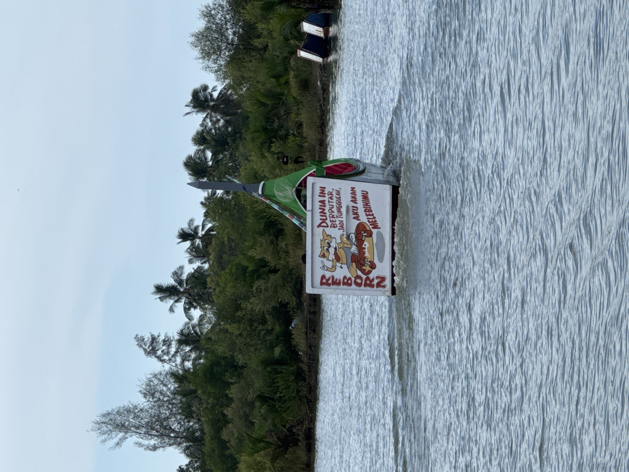 In Reudeuep, Indonesia, a boat with a white sign on its side is moving through the water. The sign features a cartoon cat character and text in Indonesian. The boat has a tall, pointed structure at its front, decorated with green and red colors. The background consists of lush green trees, including palm trees, and a cloudy sky. Two objects, possibly buoys or overturned boats, are visible in the water on the right side of the image.
