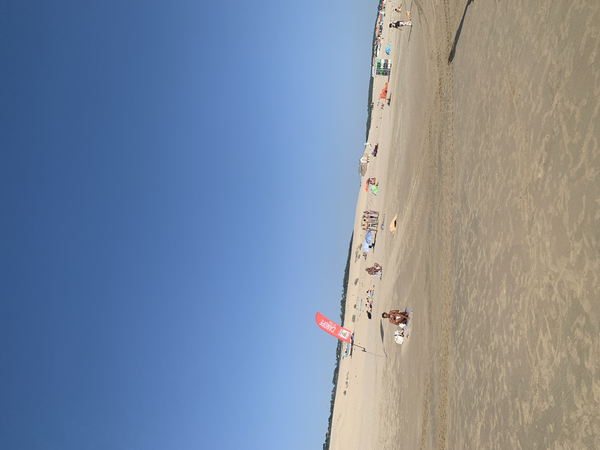 This is a photograph taken on a sunny day at Costa da Caparica, Portugal. The scene is a wide, sandy beach with a clear blue sky dominating the left side of the frame. The beach is populated with people enjoying a day out, with several individuals and small groups visible scattered across the sand. Some people are sitting or lying on towels, while others are standing. There are also beach umbrellas providing shade.
In the mid-ground and further down the beach, there are some small buildings or structures, possibly kiosks or beach facilities. A red flag with white text is planted in the sand in the foreground, with the word "CANOPE" clearly visible, along with other text that is partially obscured. The flag is attached to a pole. The sand appears to have some texture and markings, likely from wind or footsteps.
The overall impression is of a relaxed and pleasant beach atmosphere on a warm day. The bright sunlight suggests it is likely midday or early afternoon. The presence of buildings and many people indicates a popular and developed beach area.