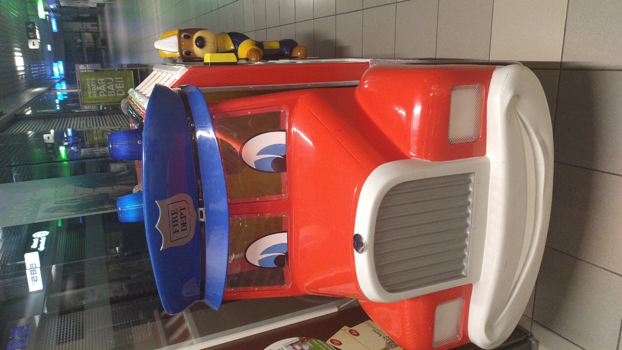 A red toy fire truck is parked indoors on a tiled floor. The truck has a blue police-style hat on top with a "FIRE DEPT" badge. The front of the truck features cartoonish eyes and a smiling bumper. To the right of the truck, a cartoon dog wearing a yellow helmet and a blue uniform is attached to the side. In the background, there are signs with text in Latvian, including "PIESĀKIET REDZES PĀR BAU DE!!" and a sign indicating a disabled person icon. The scene appears to be in a public indoor space, possibly a shopping mall or a similar venue in Jelgava, Latvia.